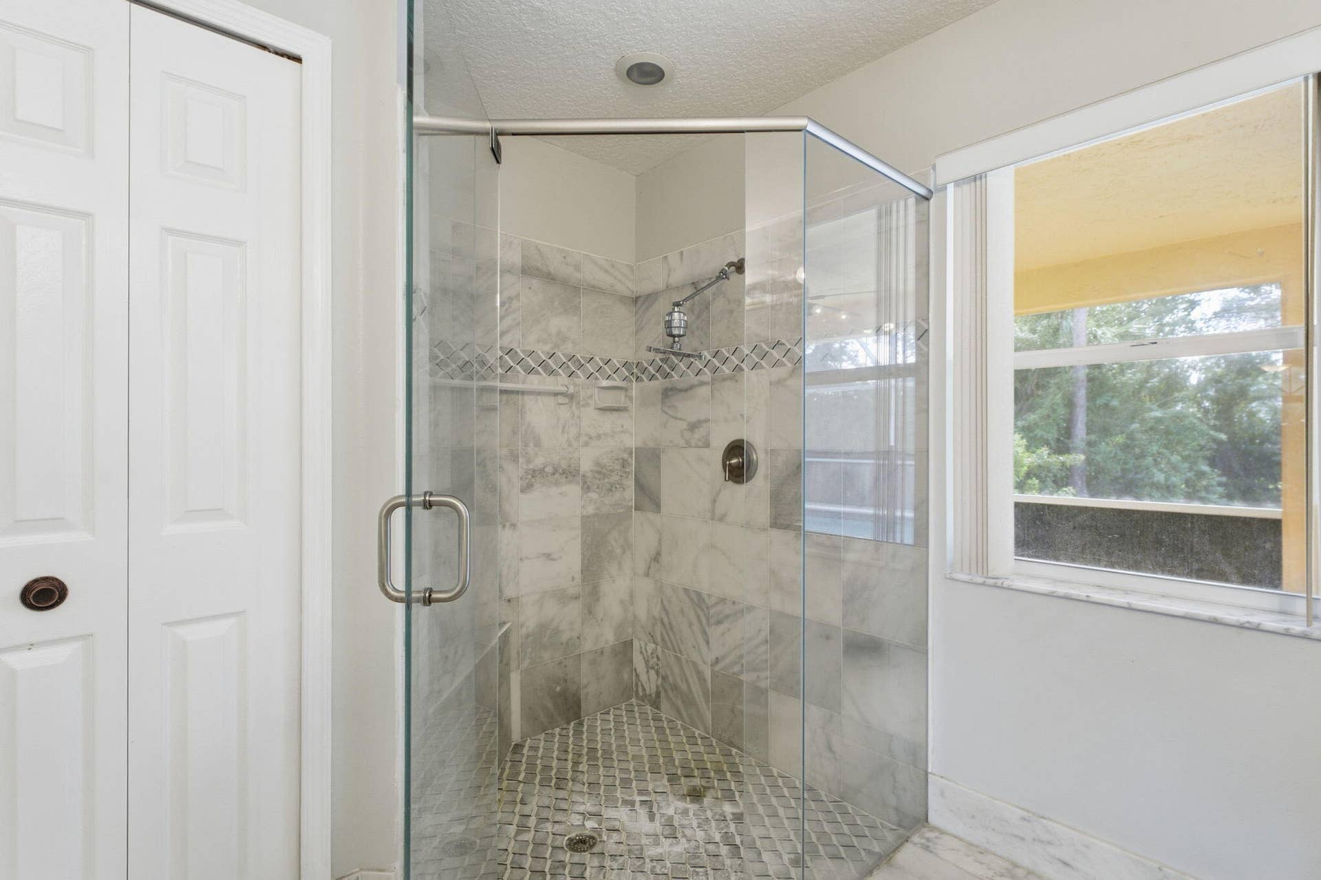 Walk-in shower featuring glass enclosure, stone-finish tile surround, accent border, and metal rainfall showerhead