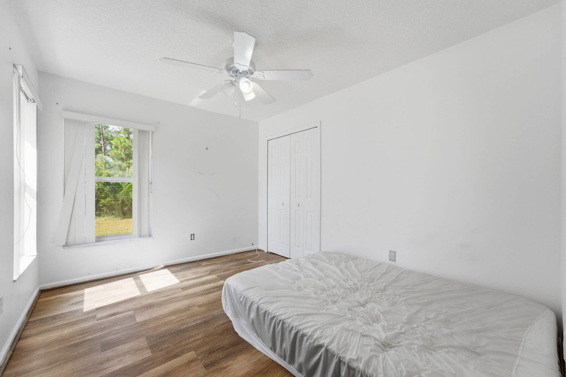 Neutral-toned interior space featuring wood-finish flooring, a white ceiling fan with integrated lighting, dual-pane windows with vertical blinds, and a double-door reach-in closet