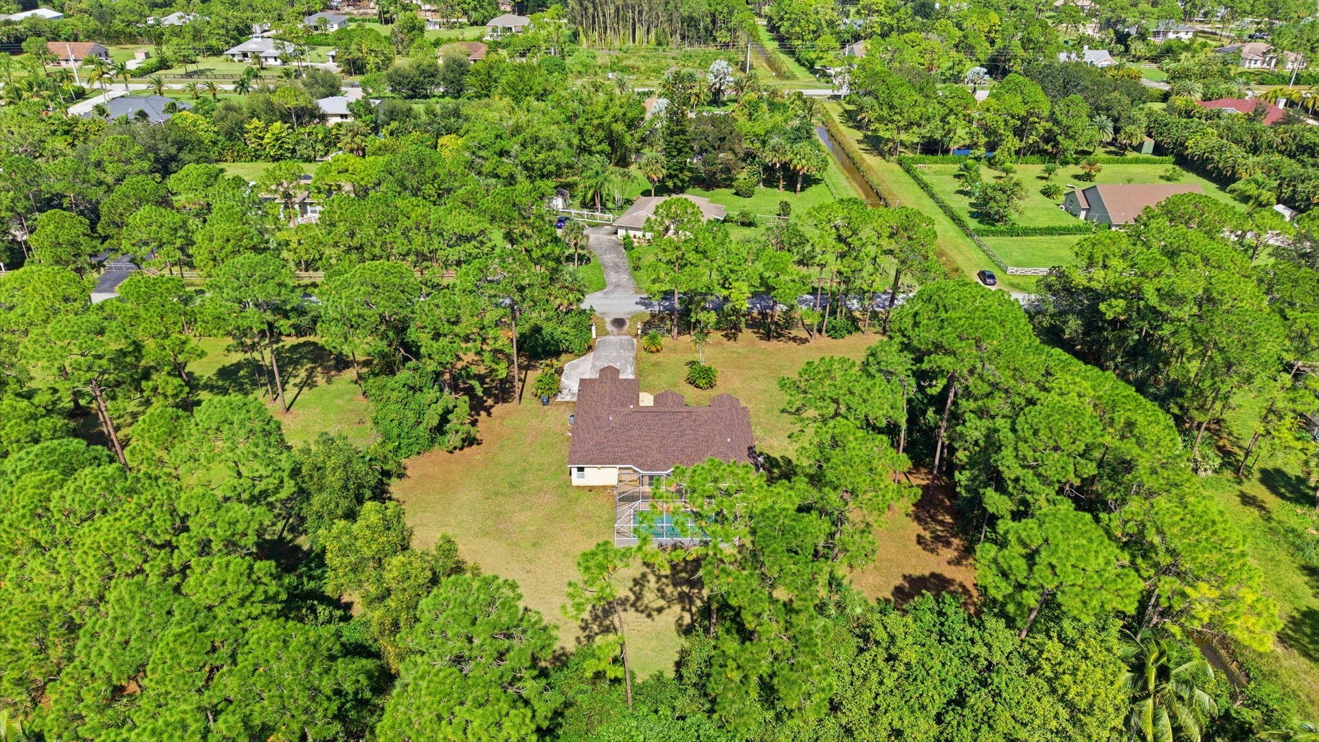 Aerial perspective featuring a single-story residence situated on a spacious lot, expansive grass lawn, mature trees, and a rectangular swimming pool with an enclosure