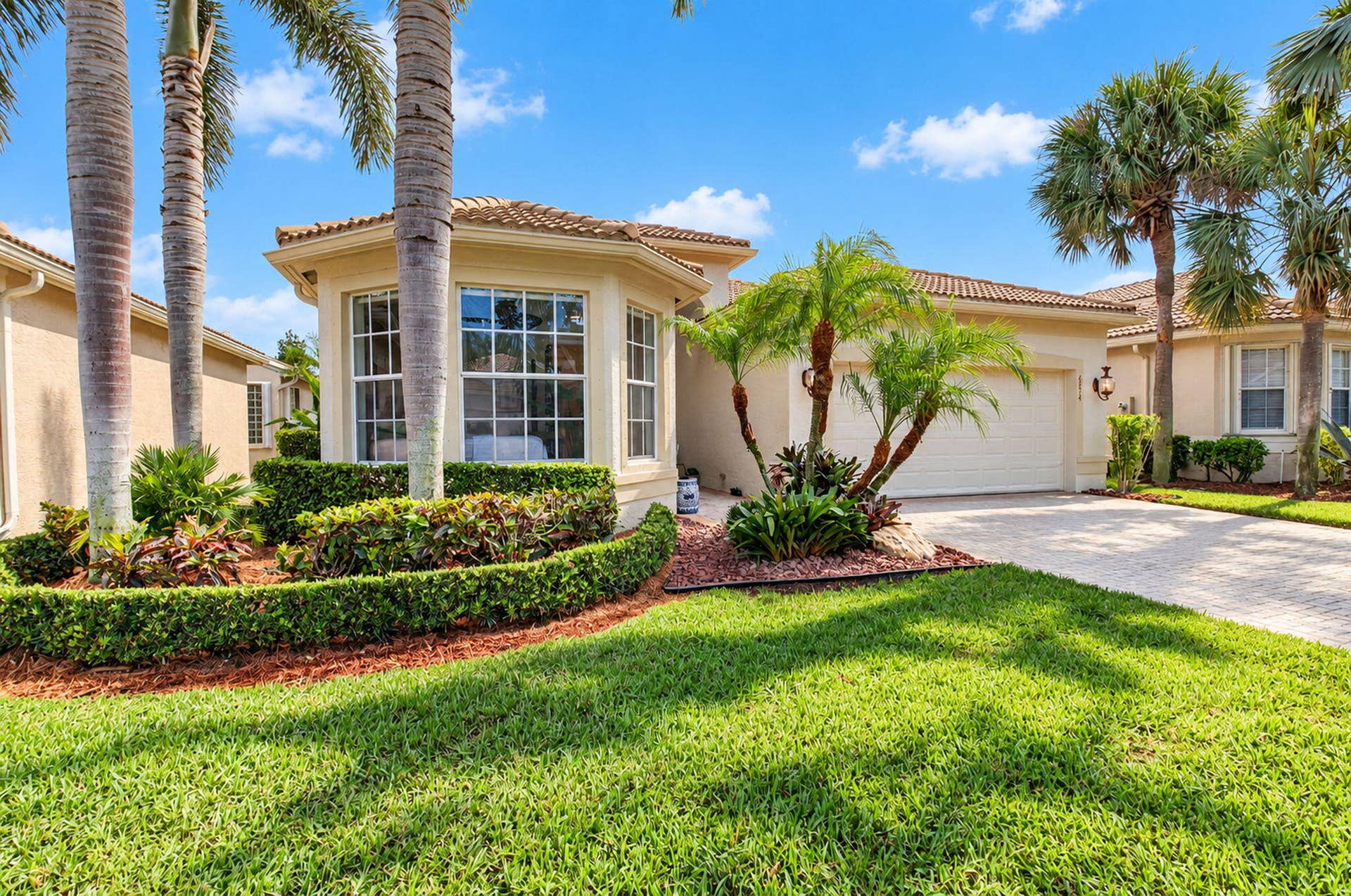 6874 Venidita Beach Drive, Delray Beach, FL 33446 Stucco exterior featuring a multi-faceted bay window, paver driveway, attached two-car garage, and manicured landscaping with mature palm trees