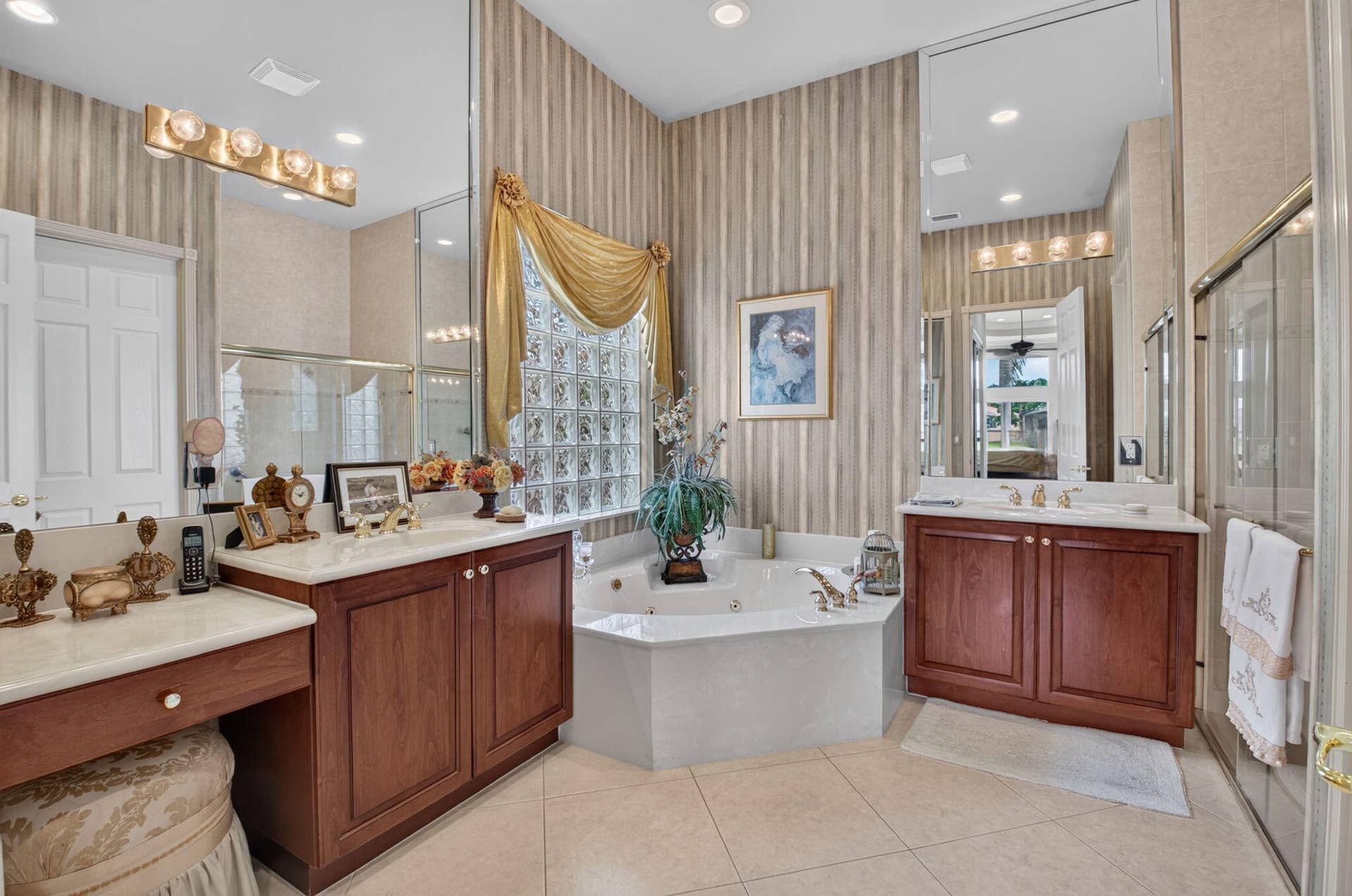 6874 Venidita Beach Drive, Delray Beach, FL 33446 Bathroom featuring dual wood-finish vanities with stone countertops, a corner jetted soaking tub, glass block accent window, and wall-to-wall mirror panels