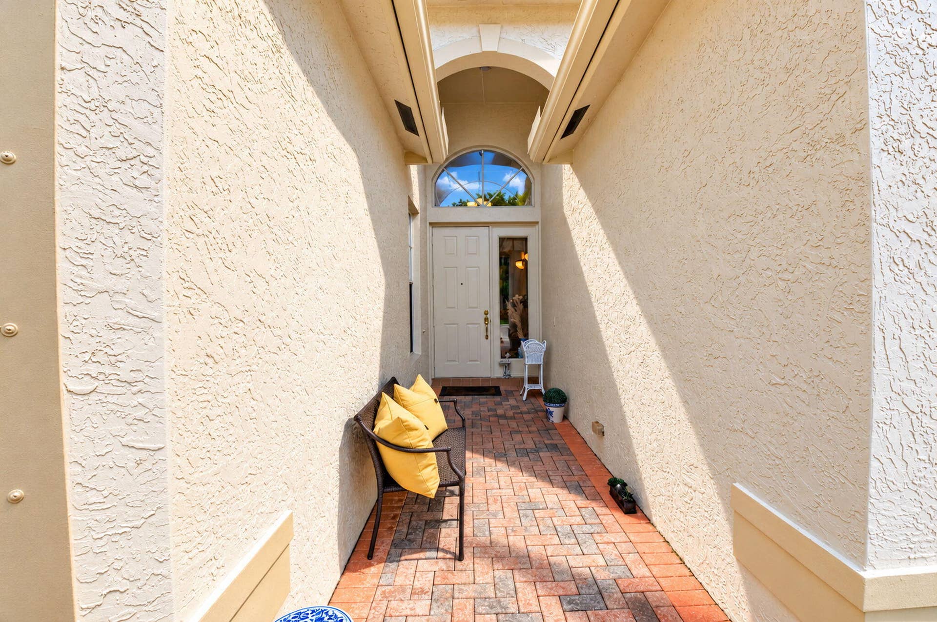 6874 Venidita Beach Drive, Delray Beach, FL 33446 Covered entryway featuring textured stucco exterior walls, brick-paver flooring, an arched entry detail, and a multi-pane door with an arched transom window