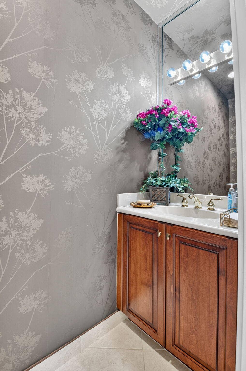 6874 Venidita Beach Drive, Delray Beach, FL 33446 Vanity featuring a stone-composite countertop, undermount sink, brass-finish faucet, and wood-toned cabinetry, accented by botanical-print wall coverings and a multi-bulb vanity light fixture