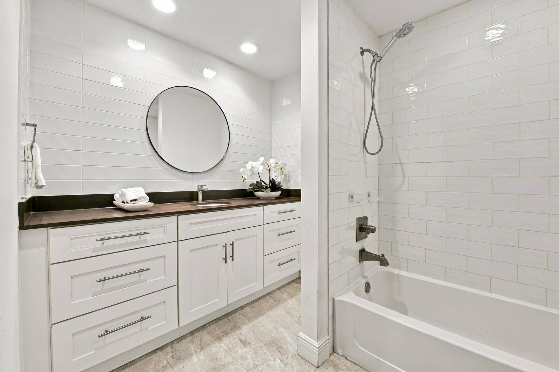 1066 NE 35TH Street, Oakland Park, FL 33334 Bathroom featuring a white shaker-style vanity with a dark countertop, round wall-mounted mirror, ceramic subway tile wall cladding, and a tub-shower combination with bronze-finish fixtures