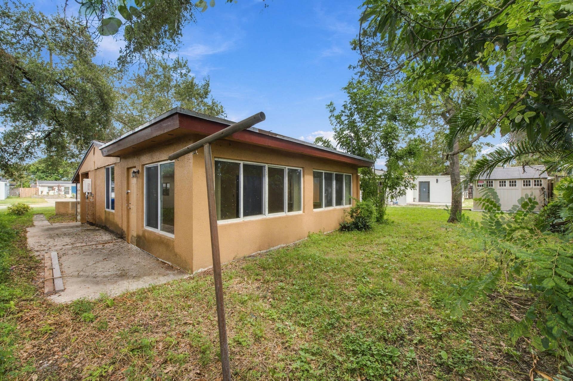6660 FREEDOM Street, Hollywood, FL 33024 Stucco exterior featuring large windows, a concrete side patio, mature trees, and a detached structure