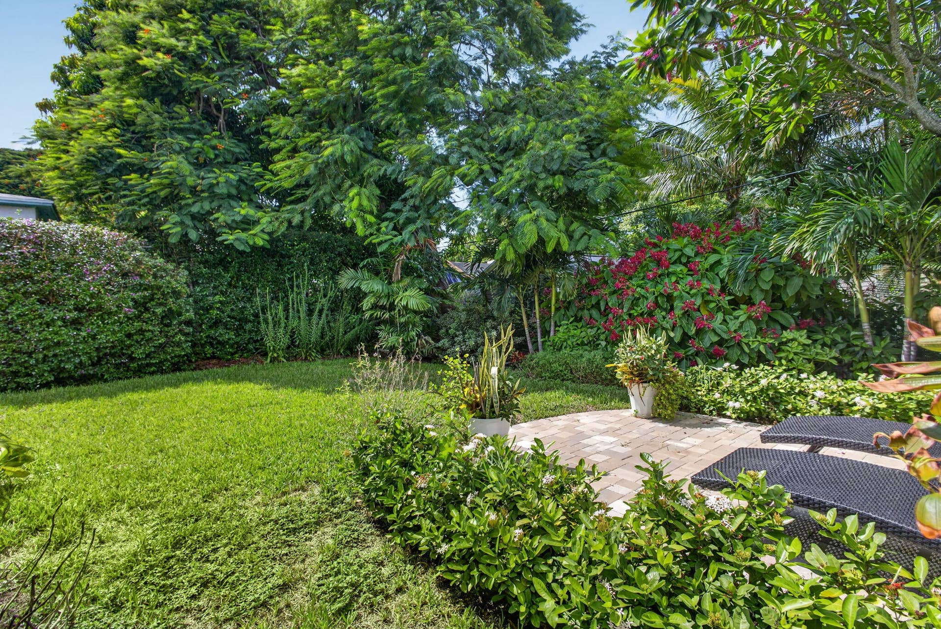 Landscaped backyard featuring a paved stone patio, mature trees, flowering shrubs, and a lawn