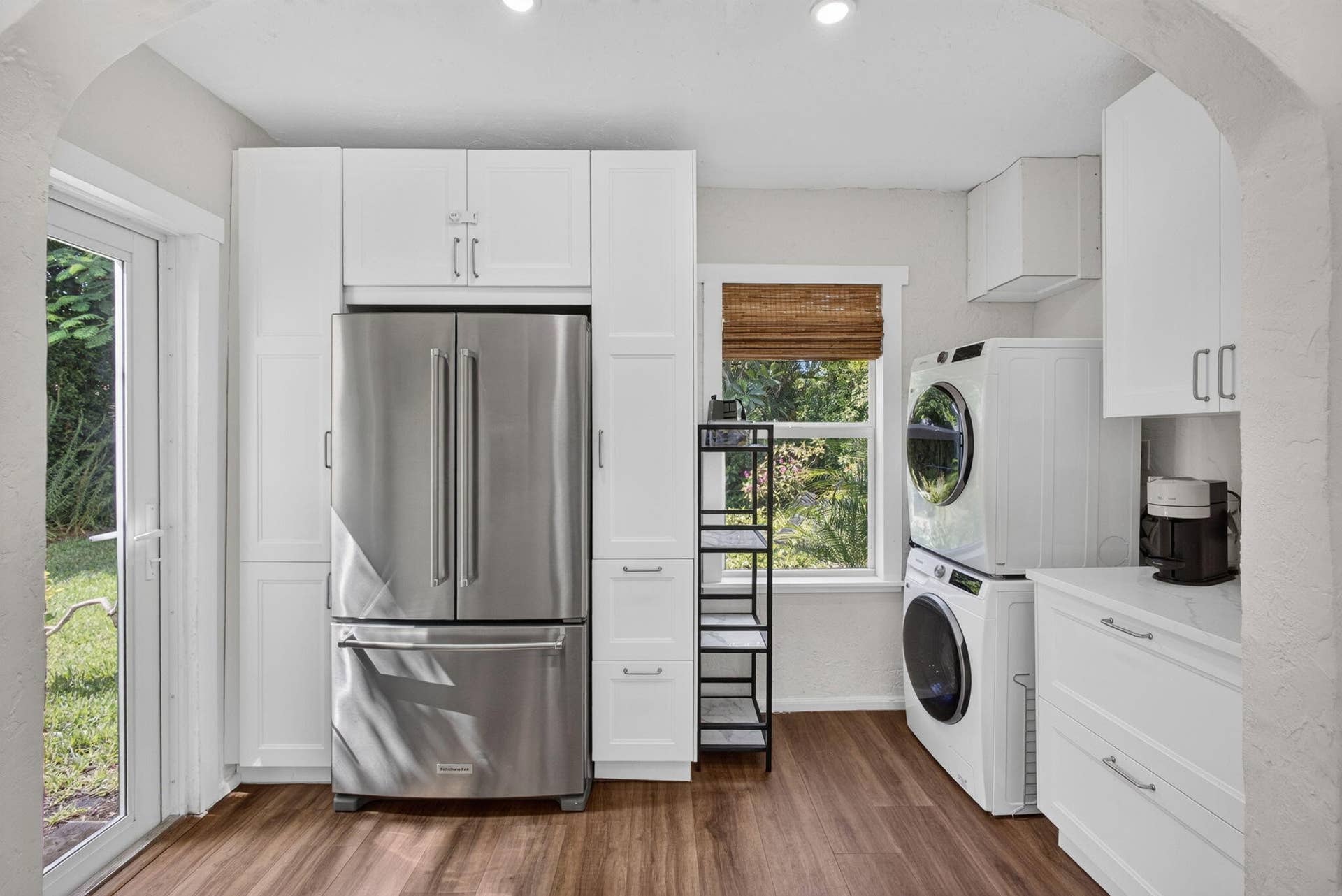 Custom white cabinetry, wood-finish flooring, stainless steel French-door refrigerator, stacked laundry unit, and a glass-paneled exterior door