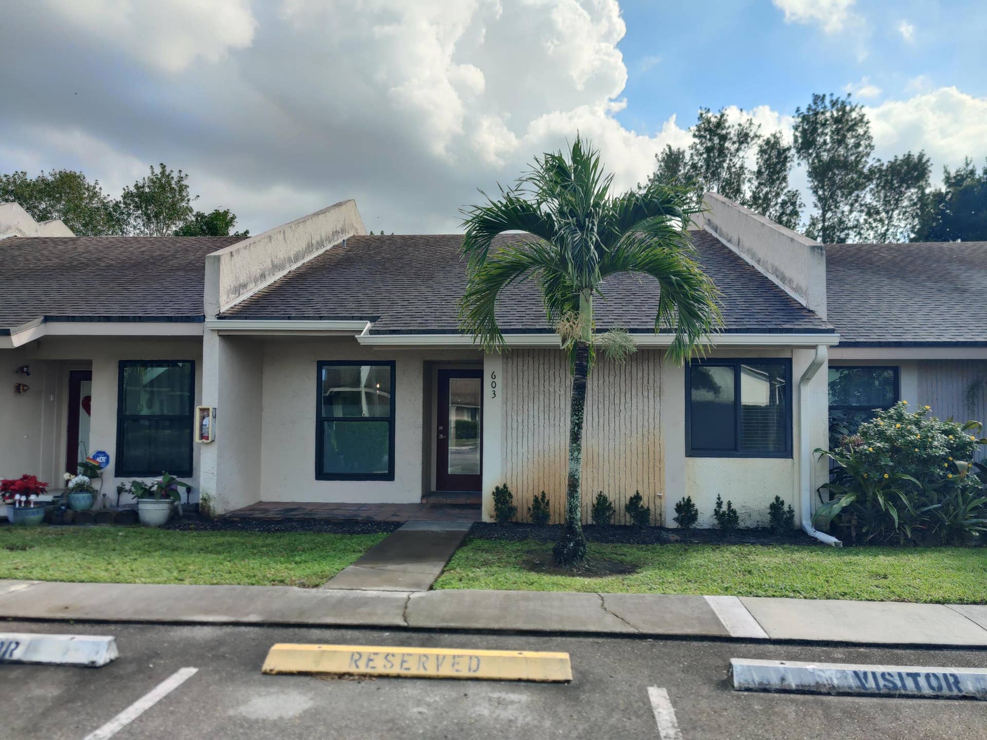 2700 S OAKLAND FOREST Drive, #603, Oakland Park, FL 33309 Single-story residence featuring a gabled roofline, stucco exterior, integrated vertical siding detail, and a prominent palm tree within the front landscape