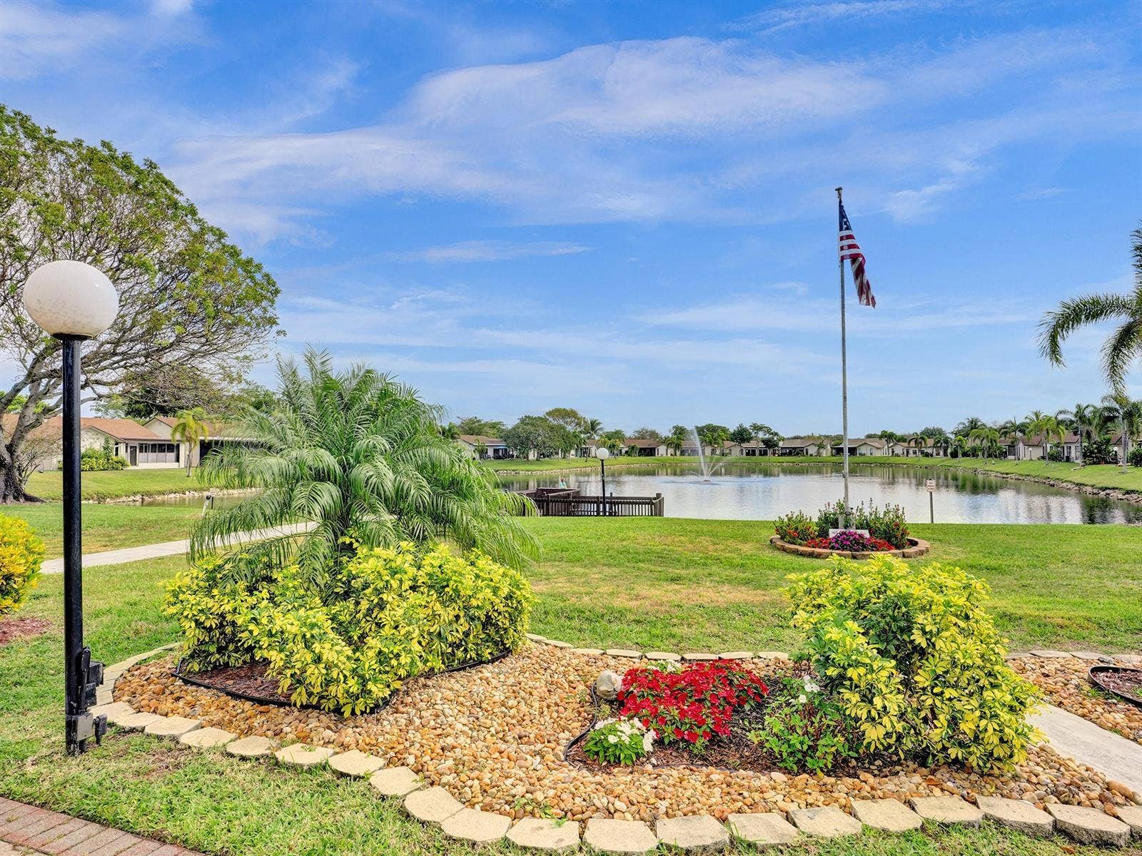 2079 SW 16TH Circle, Deerfield Beach, FL 33442 Landscaped grounds featuring a stone-lined garden bed, mature greenery, a concrete walkway, and a pond with a fountain
