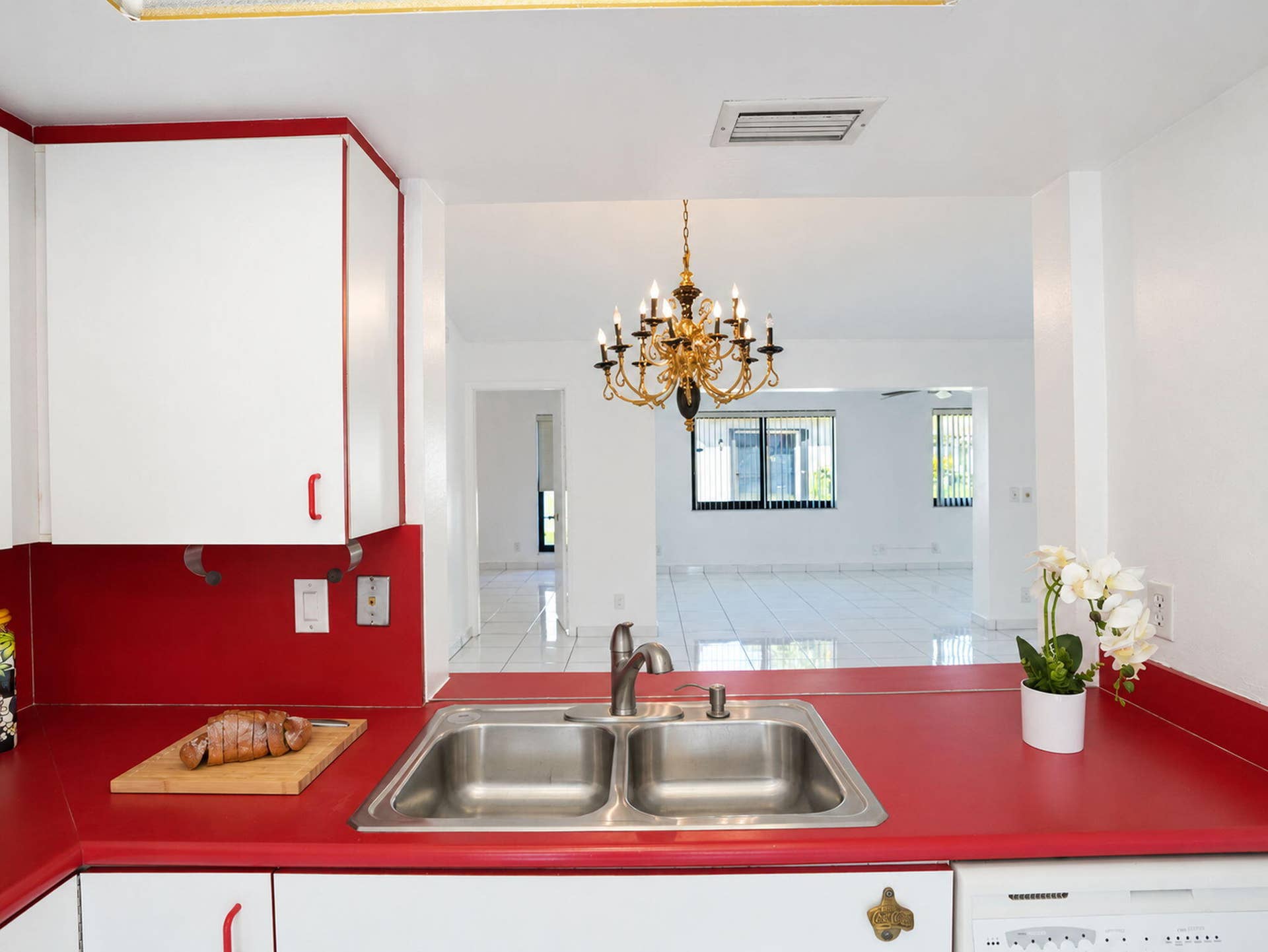2079 SW 16TH Circle, Deerfield Beach, FL 33442 Kitchen featuring a dual-basin stainless steel sink, solid-surface countertop, cabinetry with red accents, and a brass-finished multi-bulb chandelier