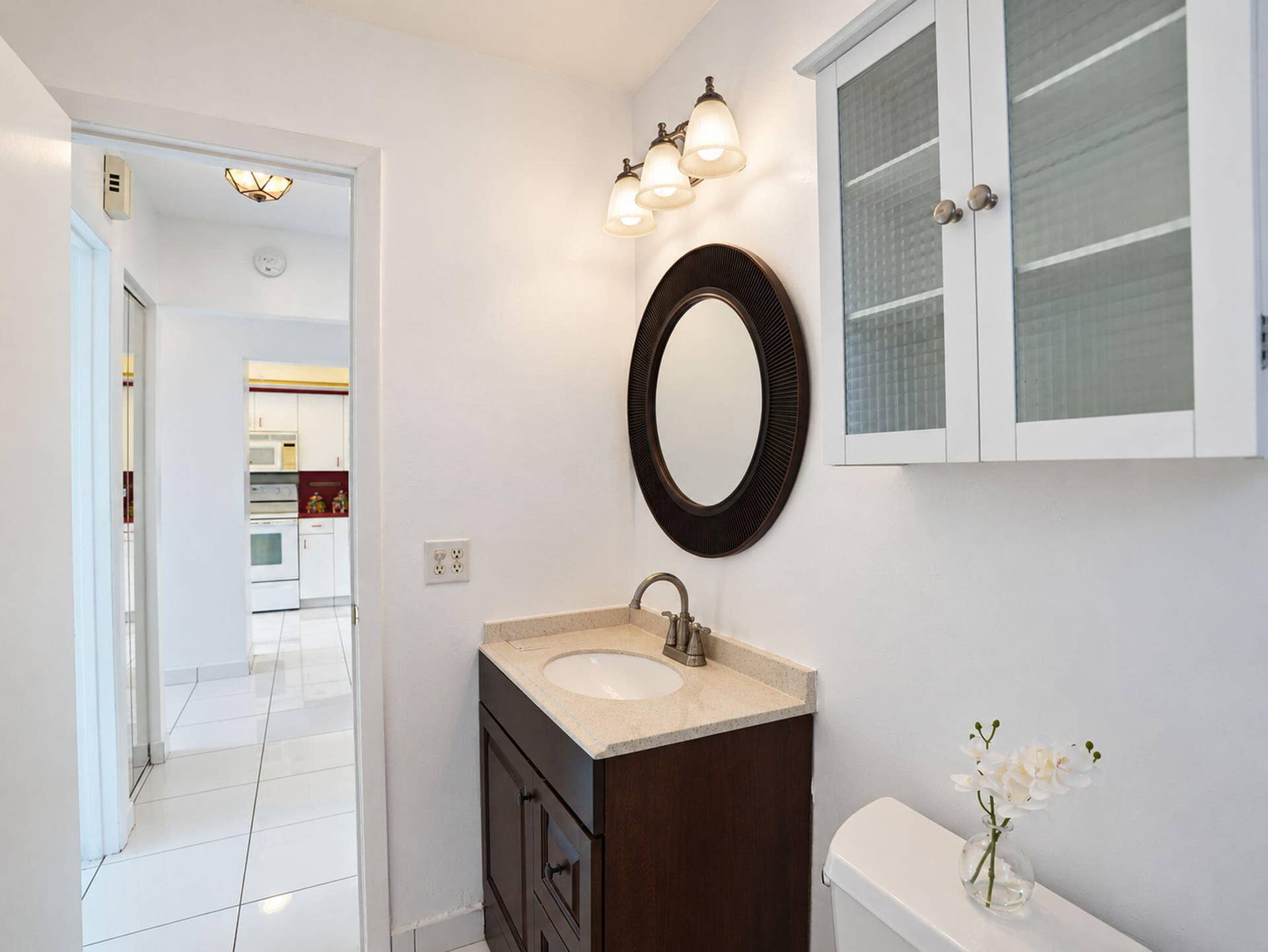 2079 SW 16TH Circle, Deerfield Beach, FL 33442 Dark wood vanity featuring an oval mirror, neutral stone countertop with an integrated basin, three-light fixture, and elevated wall-mounted cabinet with textured glass panels