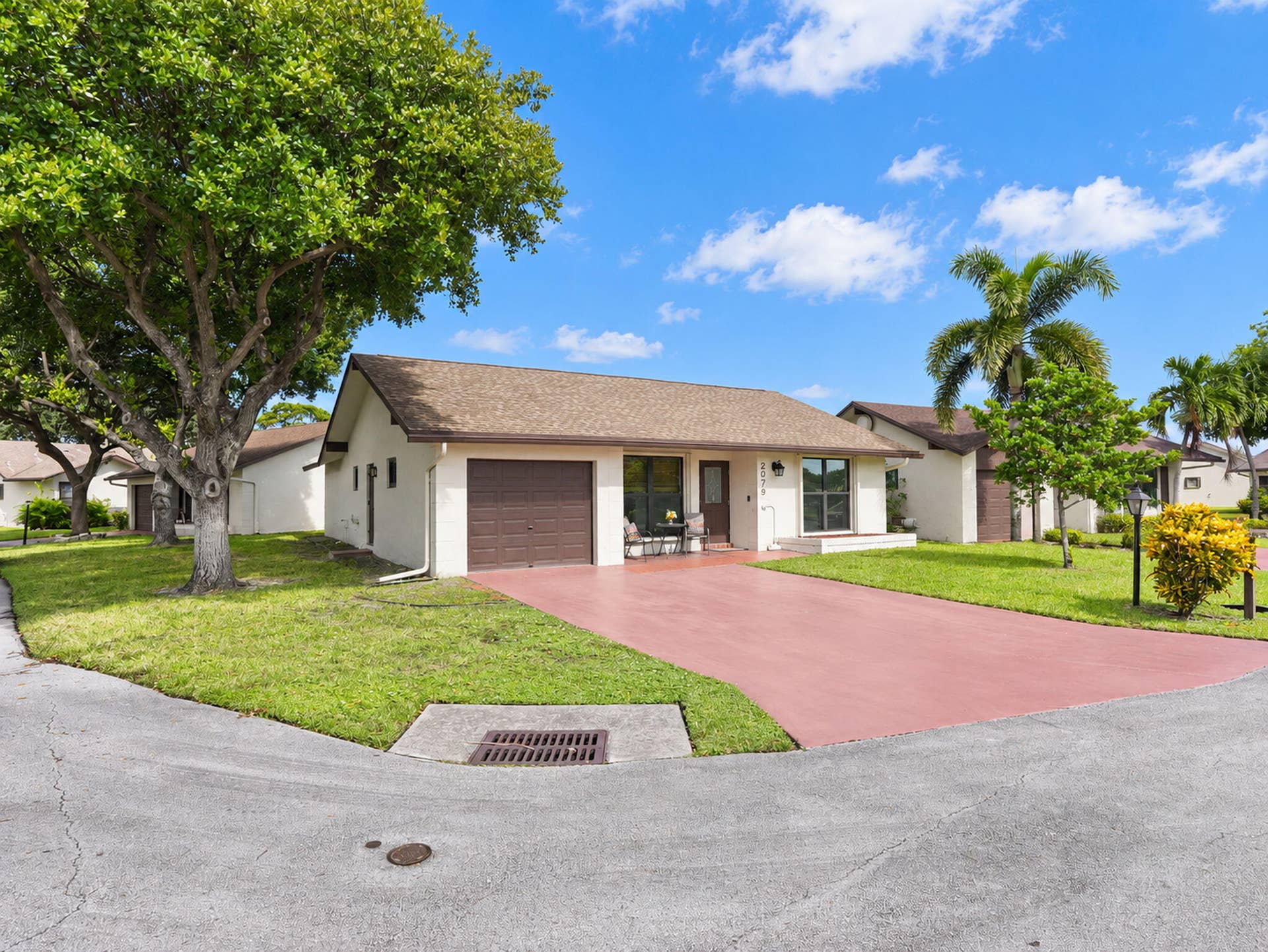 2079 SW 16TH Circle, Deerfield Beach, FL 33442 Single-story exterior featuring a shingled roof, attached one-car garage, red-toned paved driveway, and mature landscaping