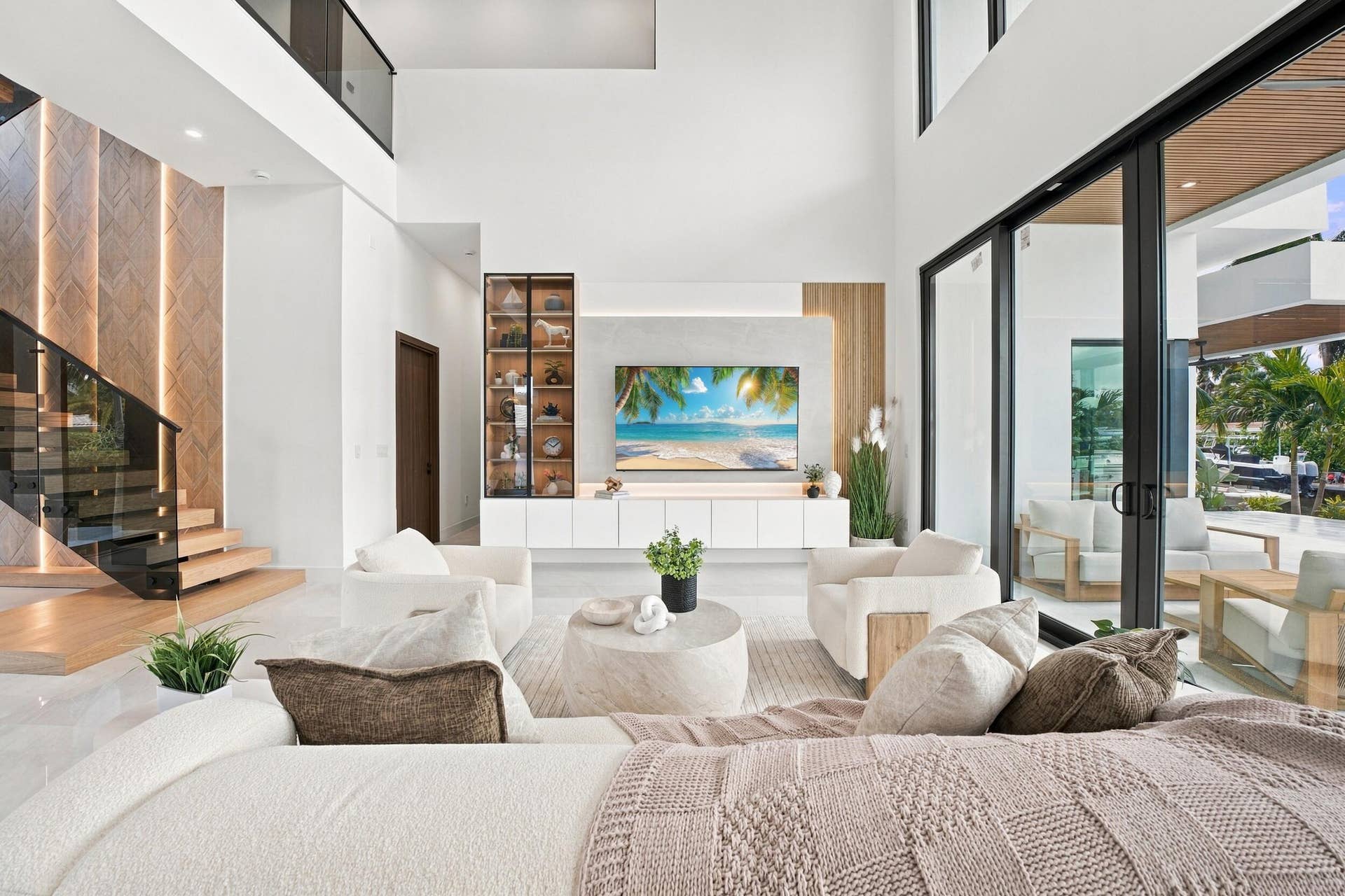 4931 NE 25TH Avenue, Lighthouse Point, FL 33064 Double-height ceilings, floating staircase with integrated vertical lighting, floor-to-ceiling glass panel doors, and custom built-in cabinetry featuring wood accents and display shelving