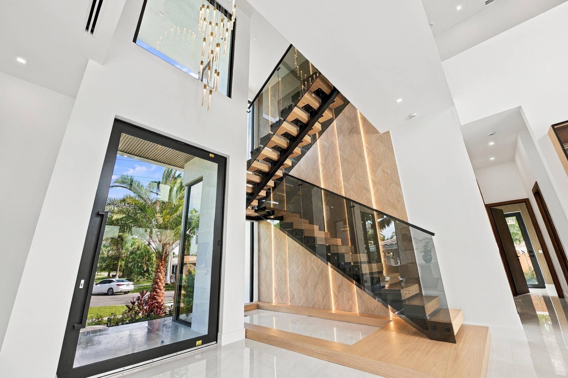 4931 NE 25TH Avenue, Lighthouse Point, FL 33064 Double-height entryway featuring a floating wood staircase with glass balustrades, large-format polished flooring, a floor-to-ceiling accent wall with integrated vertical lighting, and a prominent glass pivot entry door