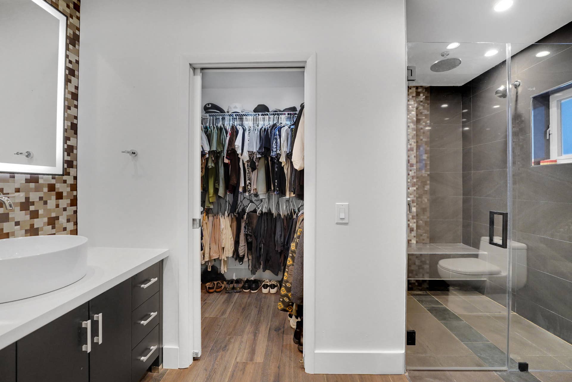 380 NW 35TH Court, Oakland Park, FL 33309 Bathroom featuring a dark wood-finish vanity with a white countertop and vessel sink, illuminated mirror, walk-in closet with built-in wire shelving, and a glass-enclosed shower with large-format tile and built-in bench