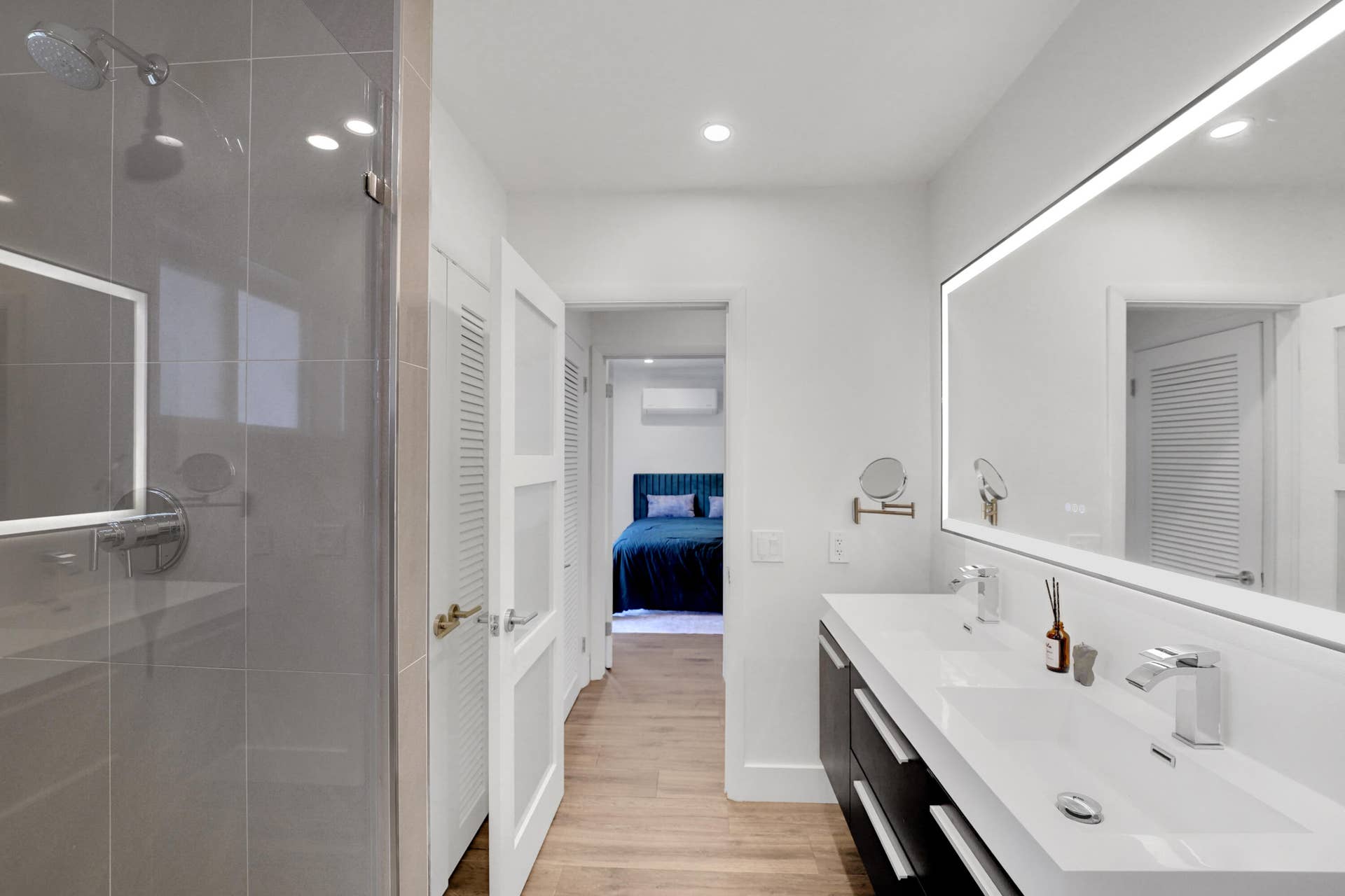 380 NW 35TH Court, Oakland Park, FL 33309 Dual-sink floating vanity with white countertops, expansive backlit mirror, glass-enclosed shower with large-format tile surround.