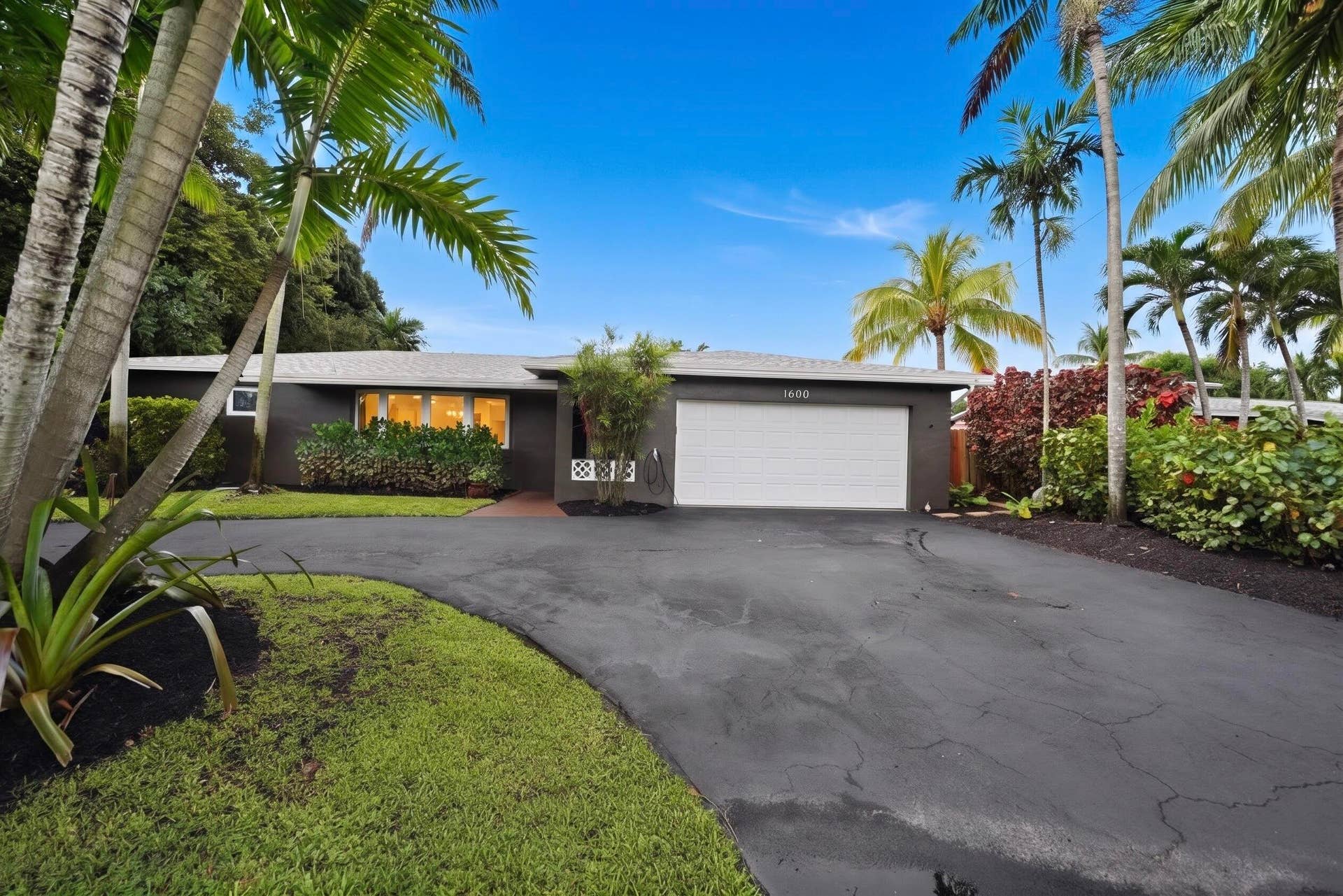 380 NW 35TH Court, Oakland Park, FL 33309 Single-story exterior featuring a dark-toned facade, an attached two-car garage, a curved asphalt driveway, and mature landscaping including palm trees