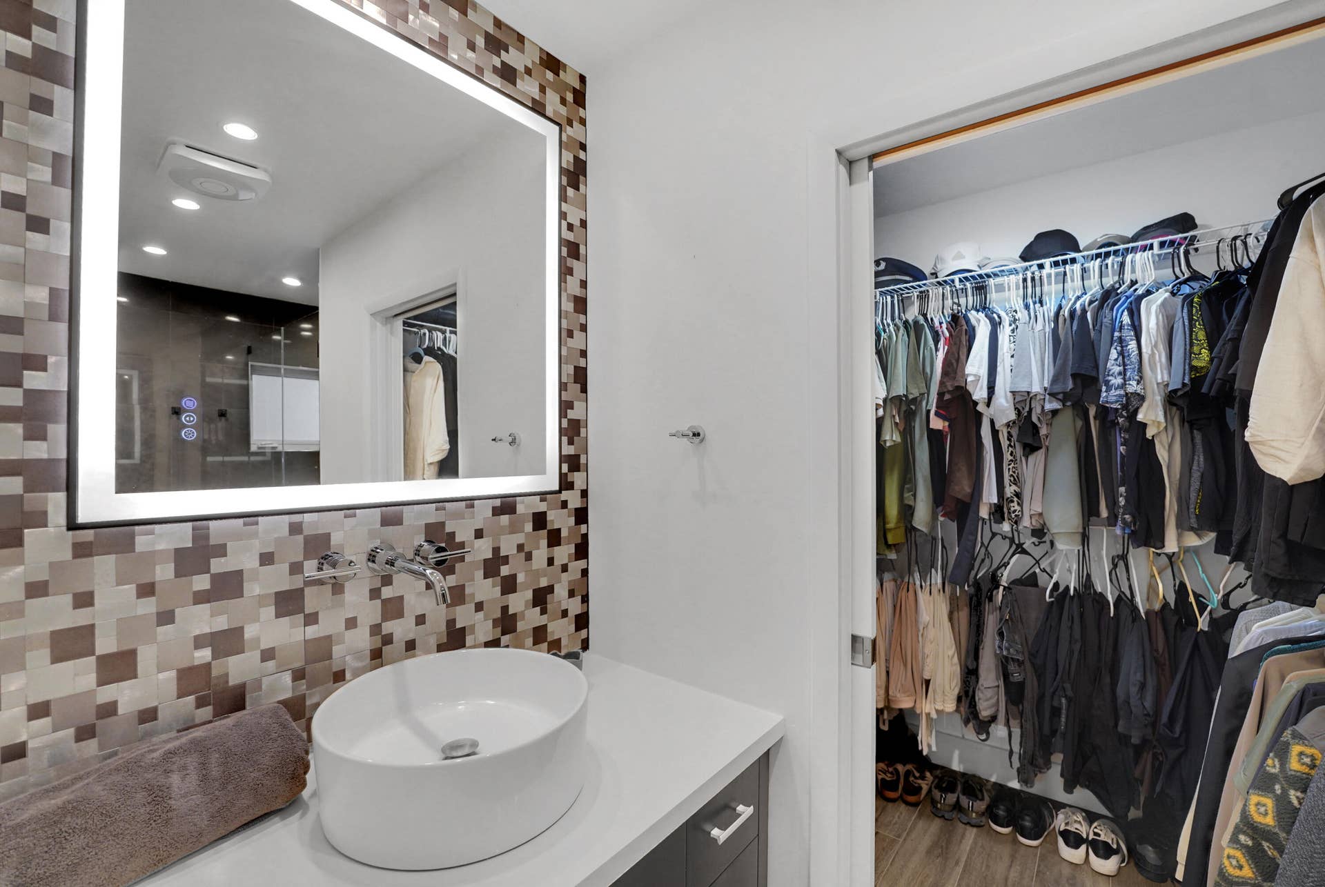 380 NW 35TH Court, Oakland Park, FL 33309 Modern bathroom featuring a rectangular vessel sink, wall-mounted faucet, illuminated mirror with integrated lighting, custom mosaic-tile backsplash, and wire-rack shelving system
