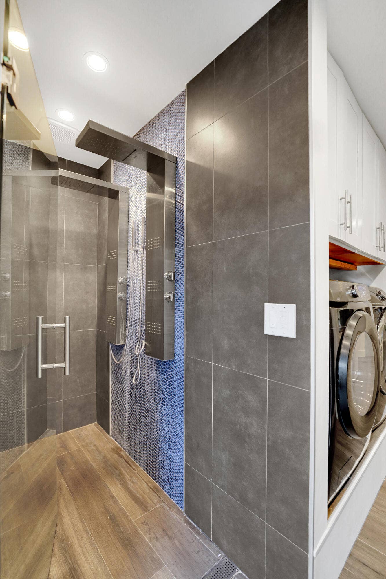 380 NW 35TH Court, Oakland Park, FL 33309 Walk-in shower featuring a metallic shower panel, blue mosaic tile accents, large-format grey wall tile, glass enclosure.