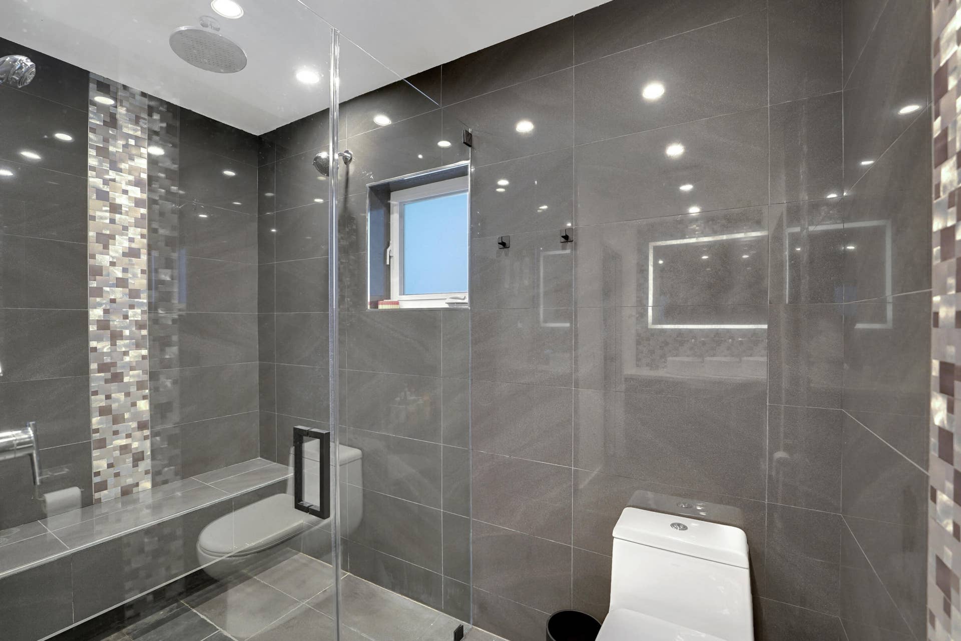 380 NW 35TH Court, Oakland Park, FL 33309 Walk-in shower featuring large-format gray tile, a vertical mosaic accent inlay, a built-in bench, a ceiling-mounted rainfall showerhead, and a frameless glass enclosure