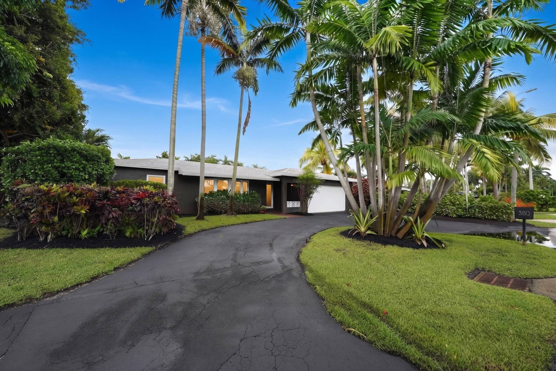380 NW 35TH Court, Oakland Park, FL 33309 Exterior facade featuring dark-toned siding, attached garage, curved asphalt driveway, and mature palm landscaping with manicured lawn