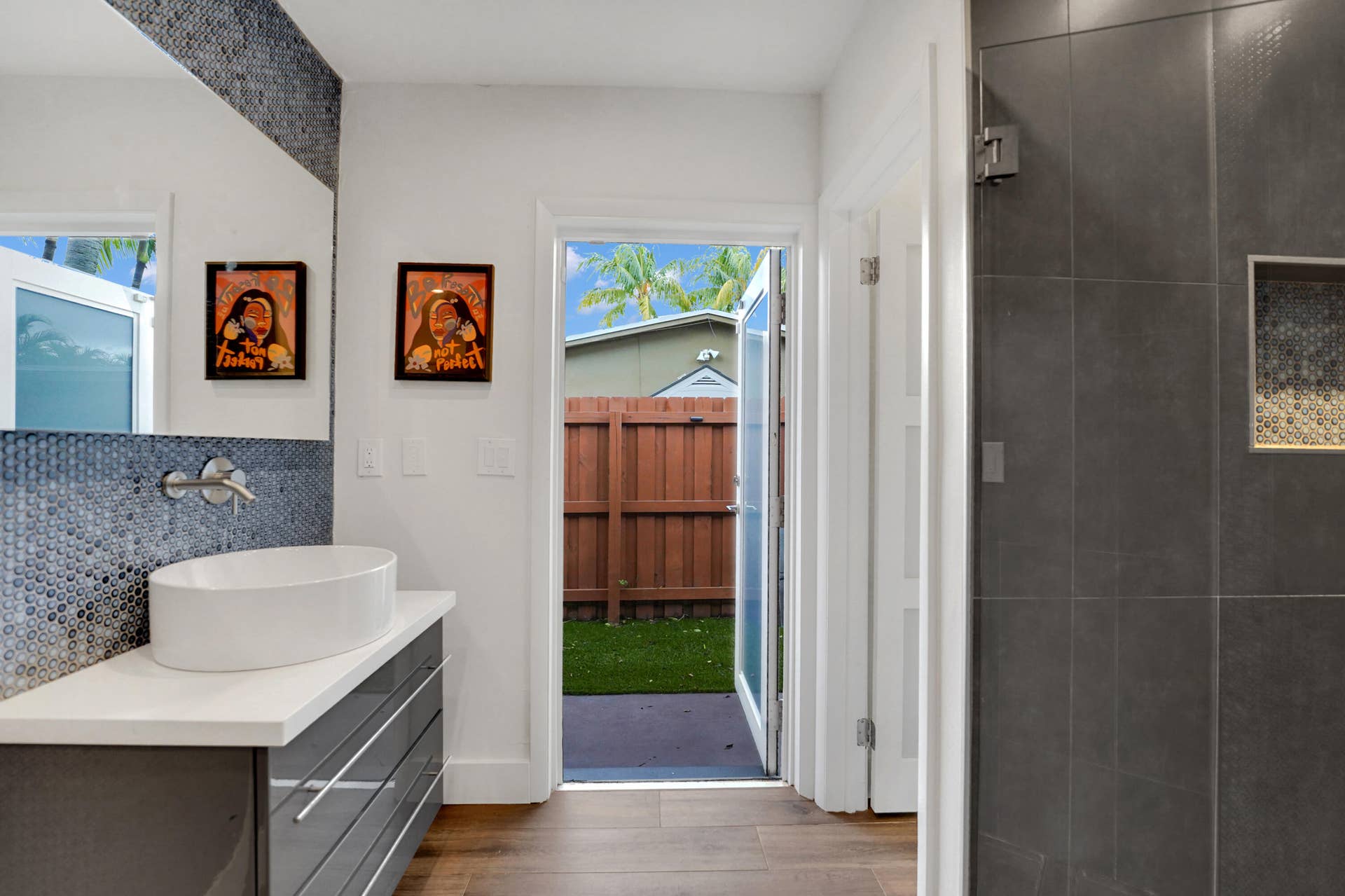 380 NW 35TH Court, Oakland Park, FL 33309 Modern floating vanity with a white vessel sink, quartz countertop, gray high-gloss cabinetry, metallic penny-round mosaic backsplash, and walk-in shower with large-format charcoal tile and a built-in recessed niche
