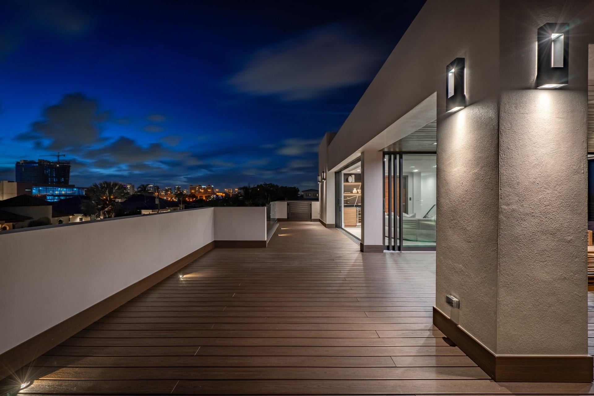 2000 Harbourview Drive, Fort Lauderdale, FL 33316 A sleek upper-level terrace illuminated by modern wall sconces, set against the dramatic glow of the evening sky. Expansive wood decking and clean architectural lines create a sophisticated outdoor retreat designed for elevated nighttime entertaining.