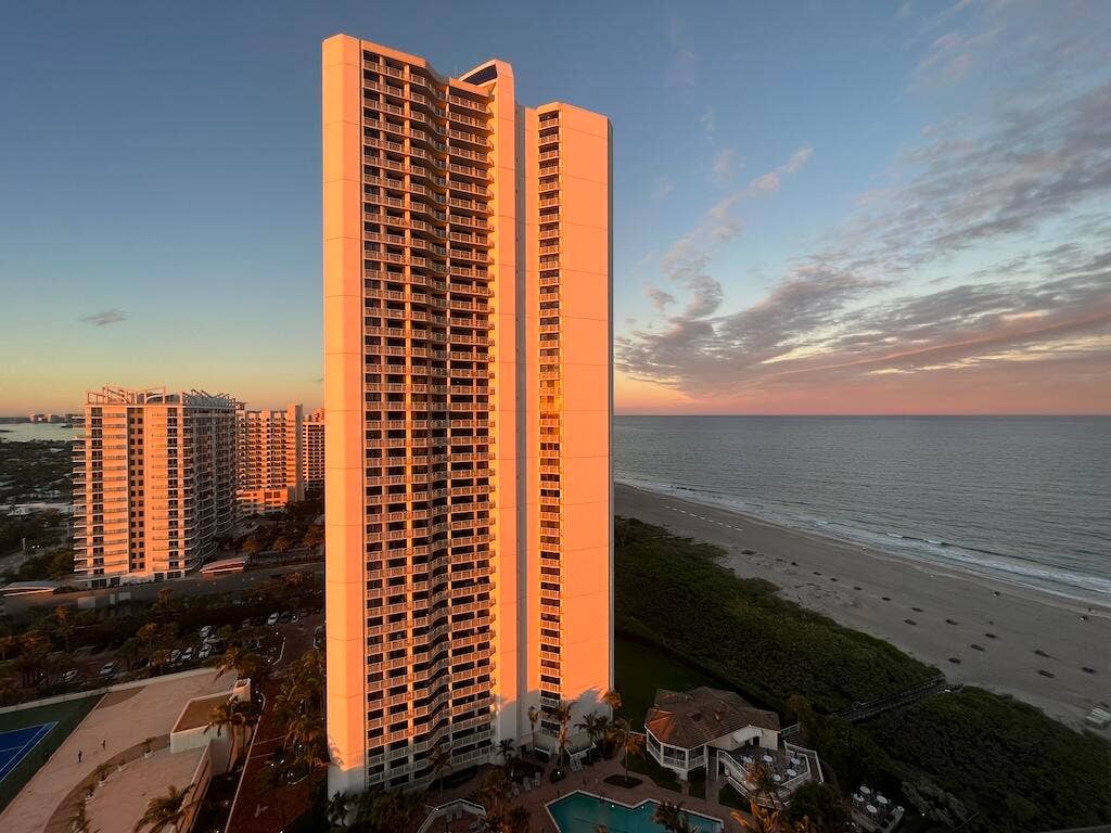 High-rise building with multiple balconies, adjacent to a sandy beach and ocean, featuring a ground-level swimming pool and tennis court