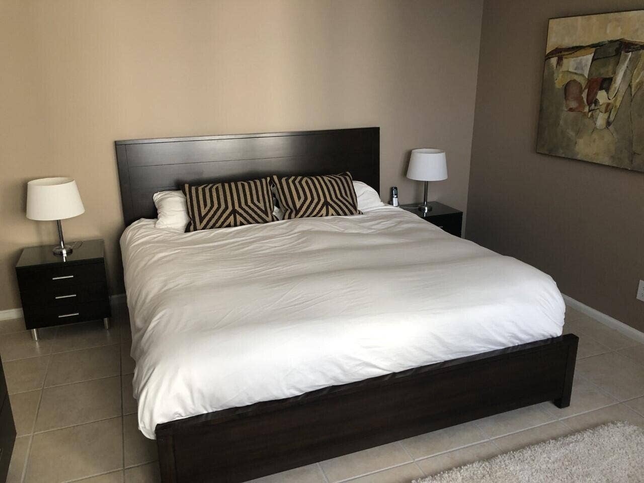Master bedroom featuring a black upholstered bed frame, dual nightstands with table lamps, neutral wall finishes, and tile flooring throughout