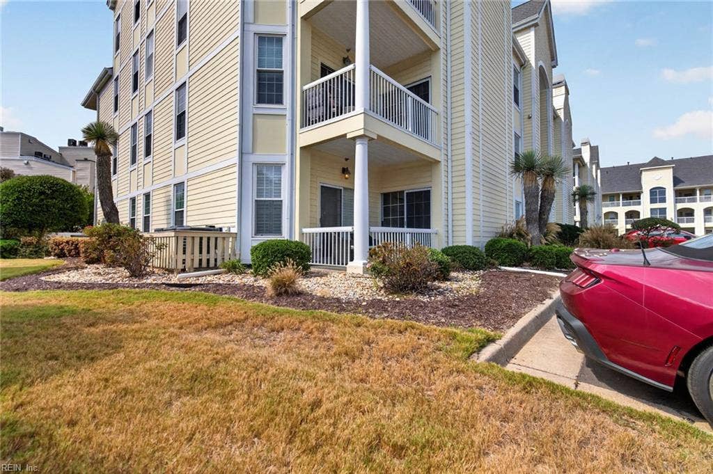 Exterior of the first-floor condo at Lesner Pointe with covered ground-level porch, sliding glass doors, and landscaped beds below two upper-floor balconies. Parking lot sits beside the building. Chic's Beach, Virginia Beach. 23451.
