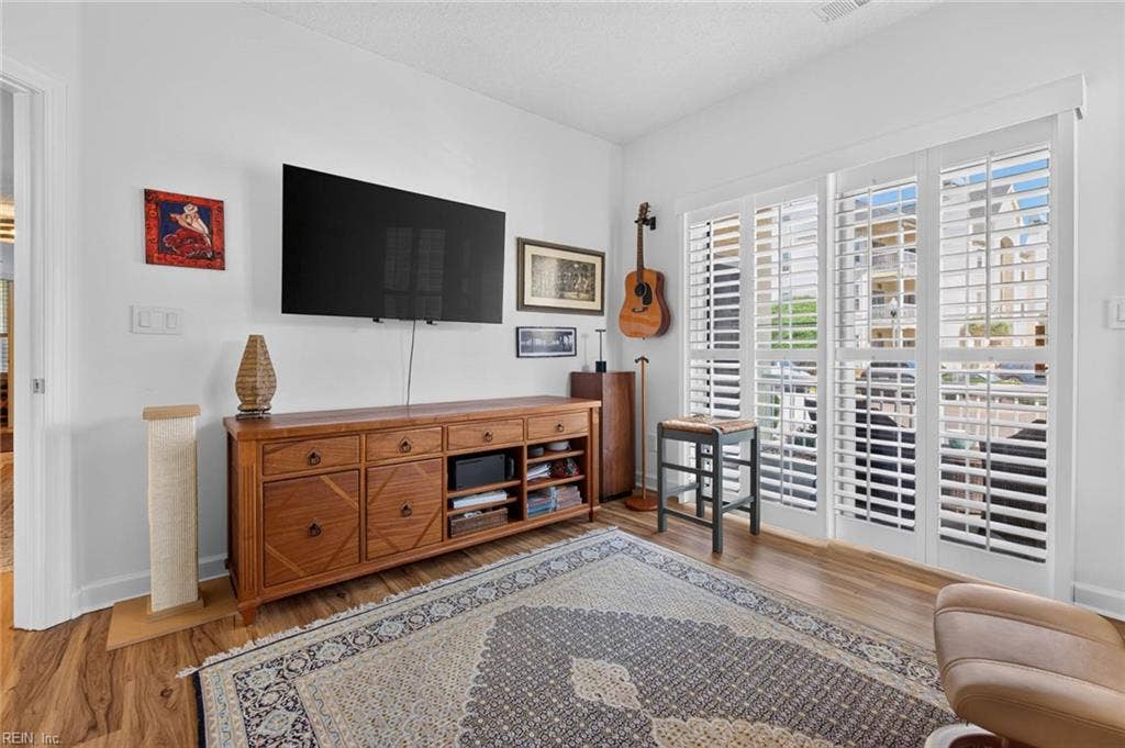 Den with sliding glass door to the covered porch, plantation shutters, ceiling fan, and LVP flooring. Shown with a wall-mounted TV and media console. Flexible third bedroom or home office space in this first-floor Lesner Pointe condo, Chic's Beach, Virginia Beach.