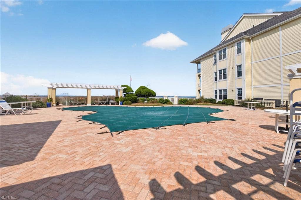 Brick paver pool deck with pergola, lounge chairs, and tables beside the Lesner Pointe pool in Chic's Beach, Virginia Beach. Dunes and the Chesapeake Bay sit just beyond the fence. Pool shown with winter cover. 23451.