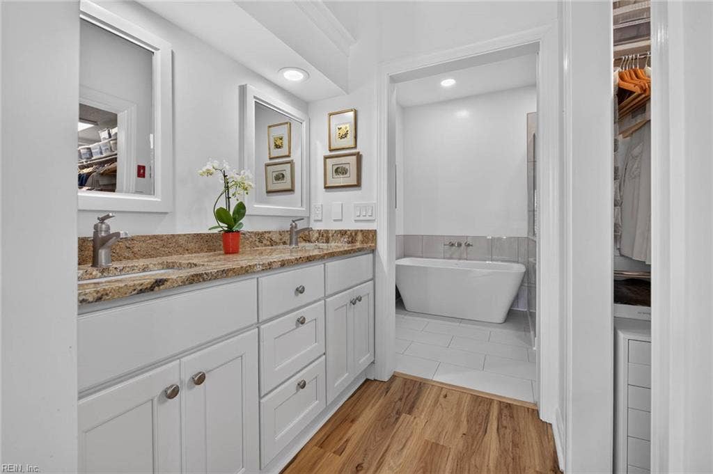 Primary bathroom with dual-sink vanity, granite countertop, white cabinets, and separate freestanding tub room with large-format tile floor. Walk-in closet entry at right. Gutted and renovated by current owners. Lesner Pointe, Chic's Beach, Virginia Beach.
