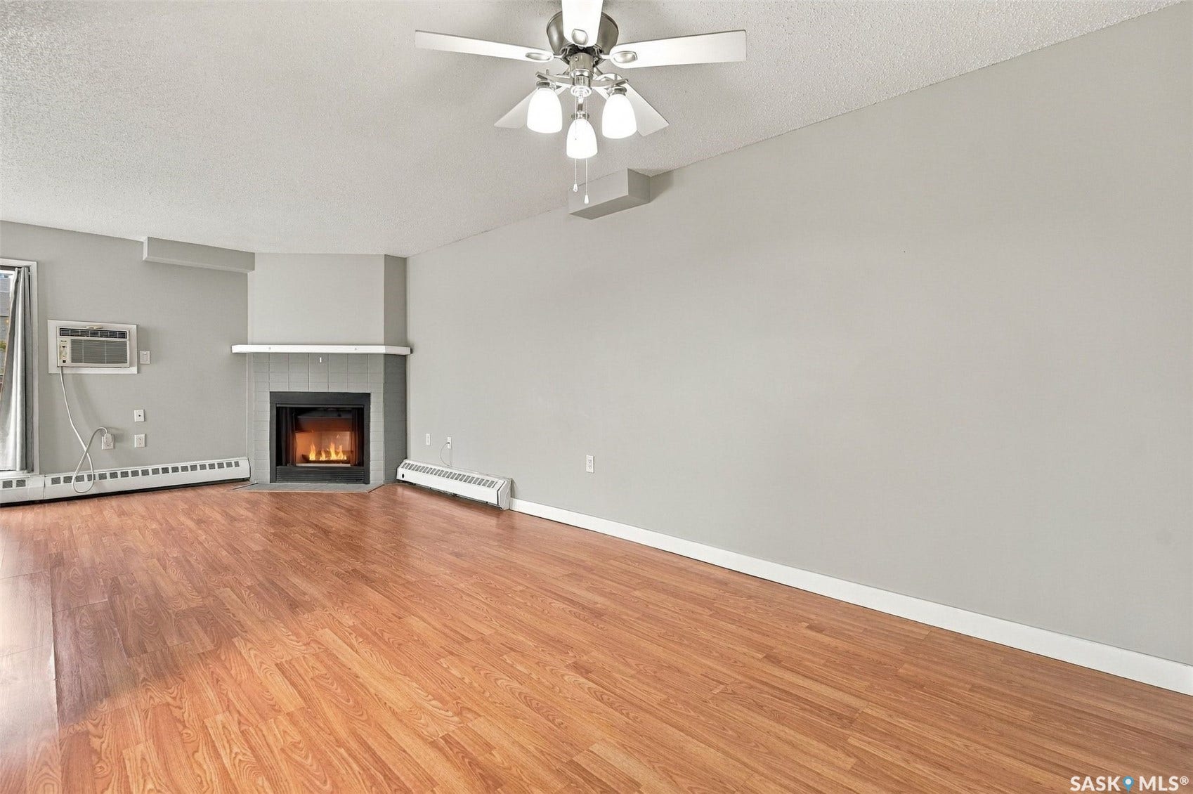 3410 Park Street, #119, Regina SK S4V 2M8 Spacious room with wood-finish flooring, a ceiling fan with integrated lighting, and a corner fireplace with a mantel