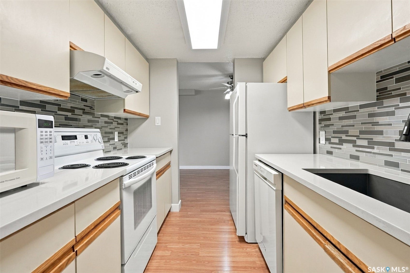 3410 Park Street, #119, Regina SK S4V 2M8 Galley kitchen featuring white appliances, light-toned cabinetry with wood trim, grey and white mosaic tile backsplash, and light countertops