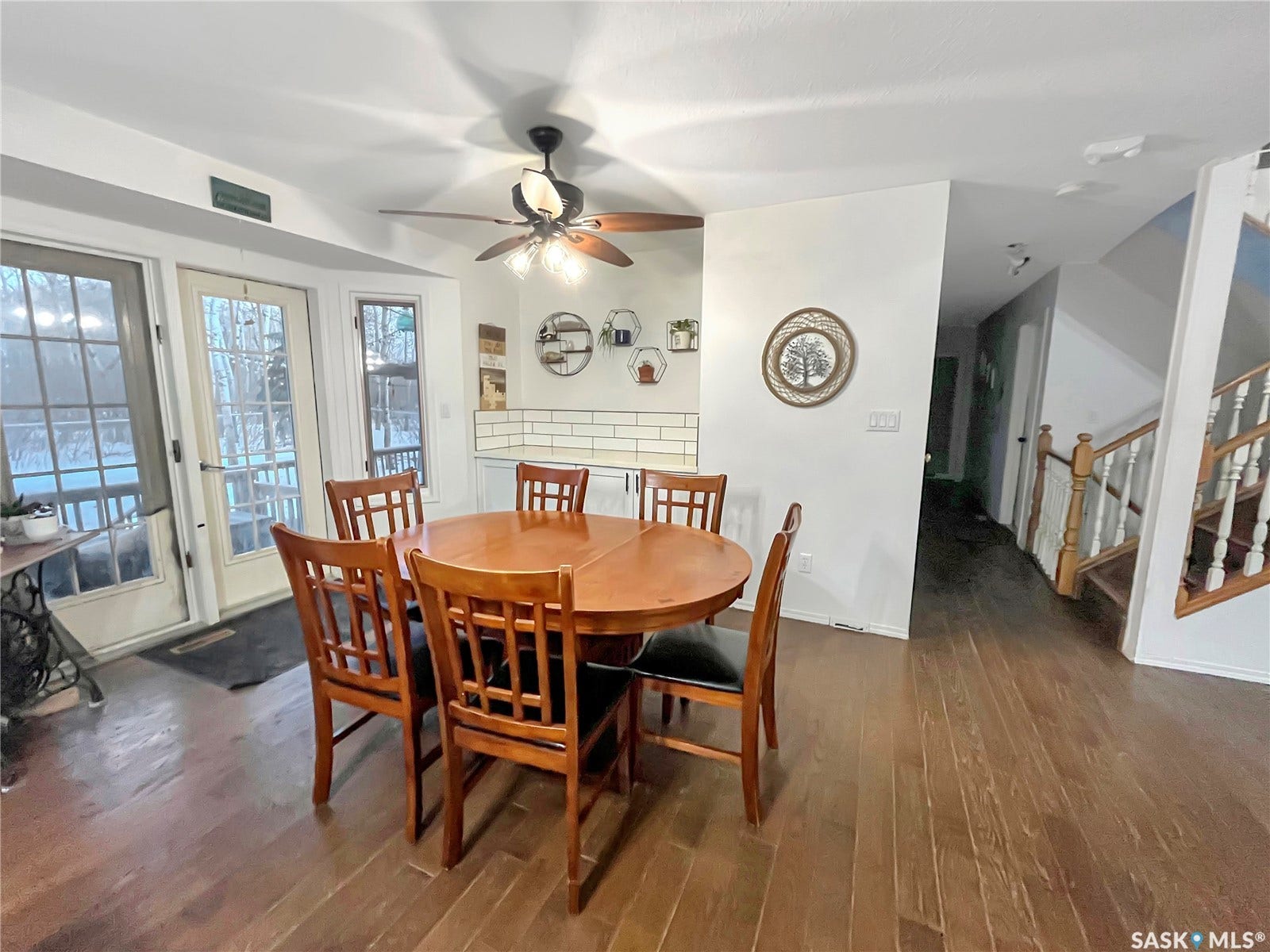 Wood-finish flooring throughout, ceiling fan with integrated lighting, two glass-paned doors providing exterior access, and a kitchen area featuring white subway tile backsplash