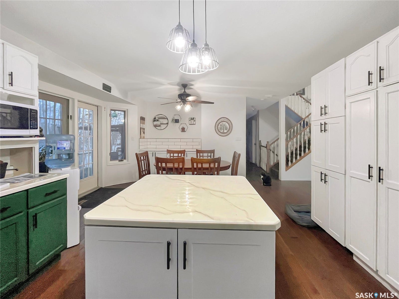 Central island with a light-toned countertop, extensive white cabinetry with dark hardware, wood-finish flooring, a dining area with a ceiling fan, and a staircase with wood newel posts and railings