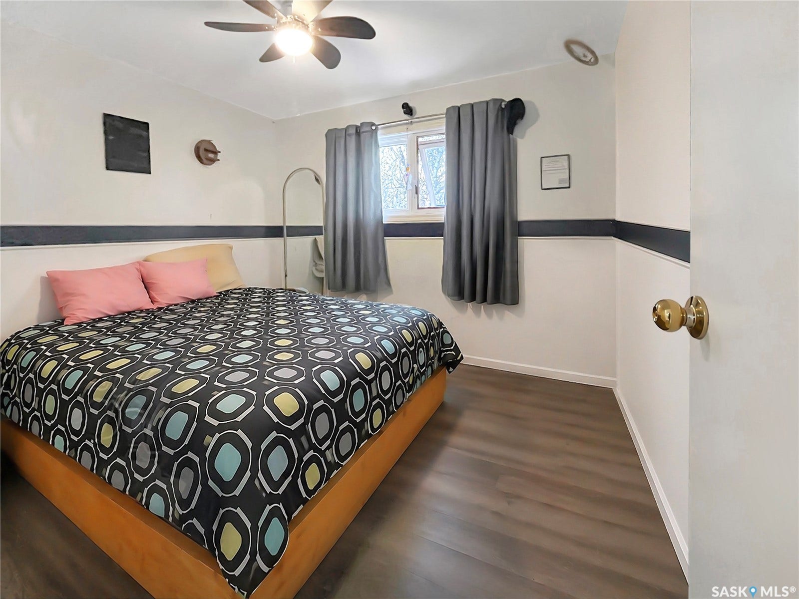 Wood-finish flooring with crisp white baseboards, ceiling fan with integrated lighting, and a single window with curtain rods