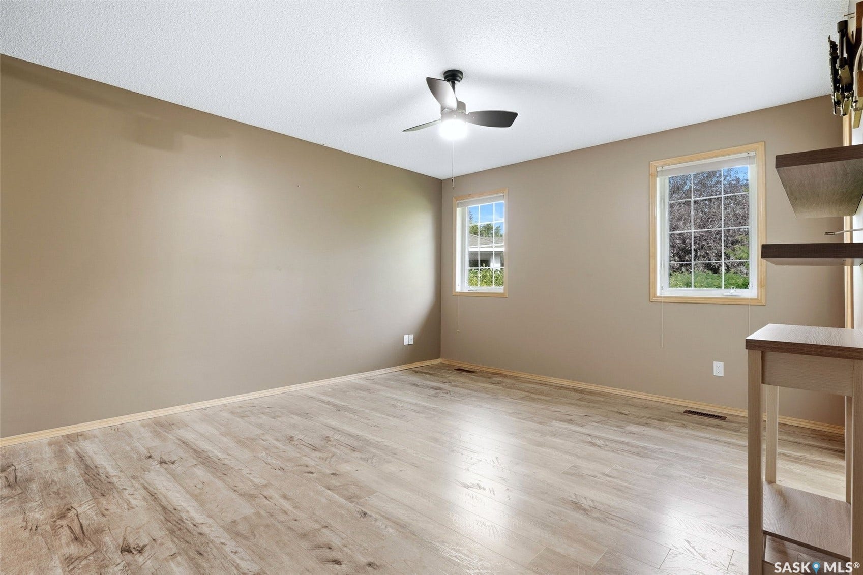 Wood-finish flooring, two windows with natural wood trim, a ceiling fan with integrated lighting, and a built-in shelving unit
