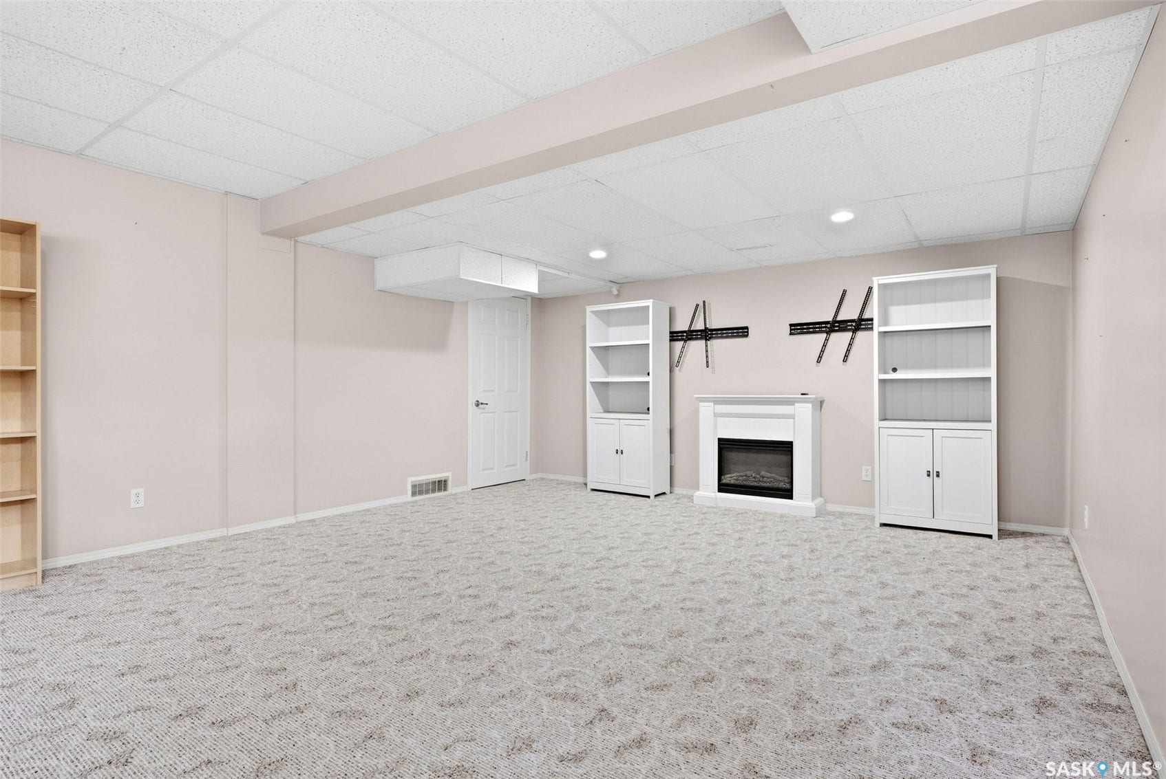 Recessed lighting illuminates a carpeted living space with a built-in electric fireplace and two white built-in shelving units