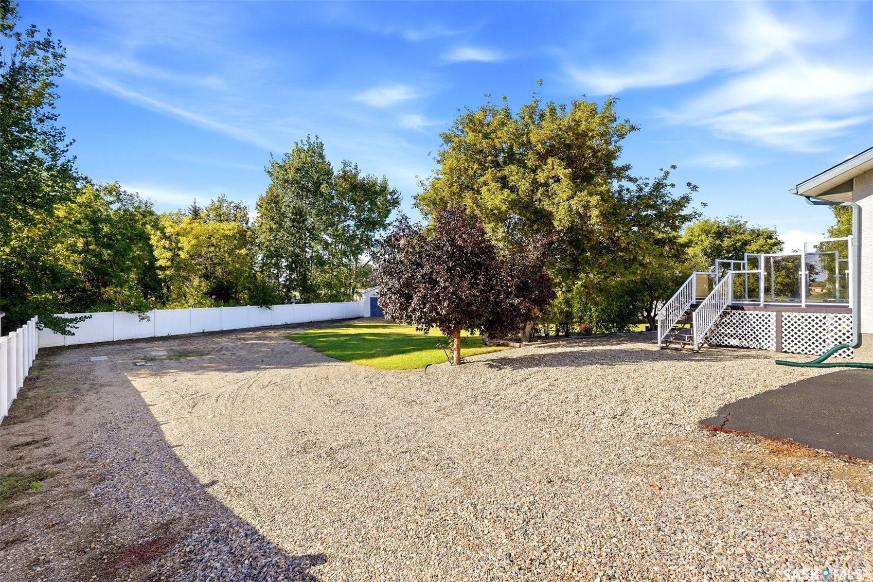 Gravel parking area with a white privacy fence, mature trees, and a raised exterior deck with glass panel railing