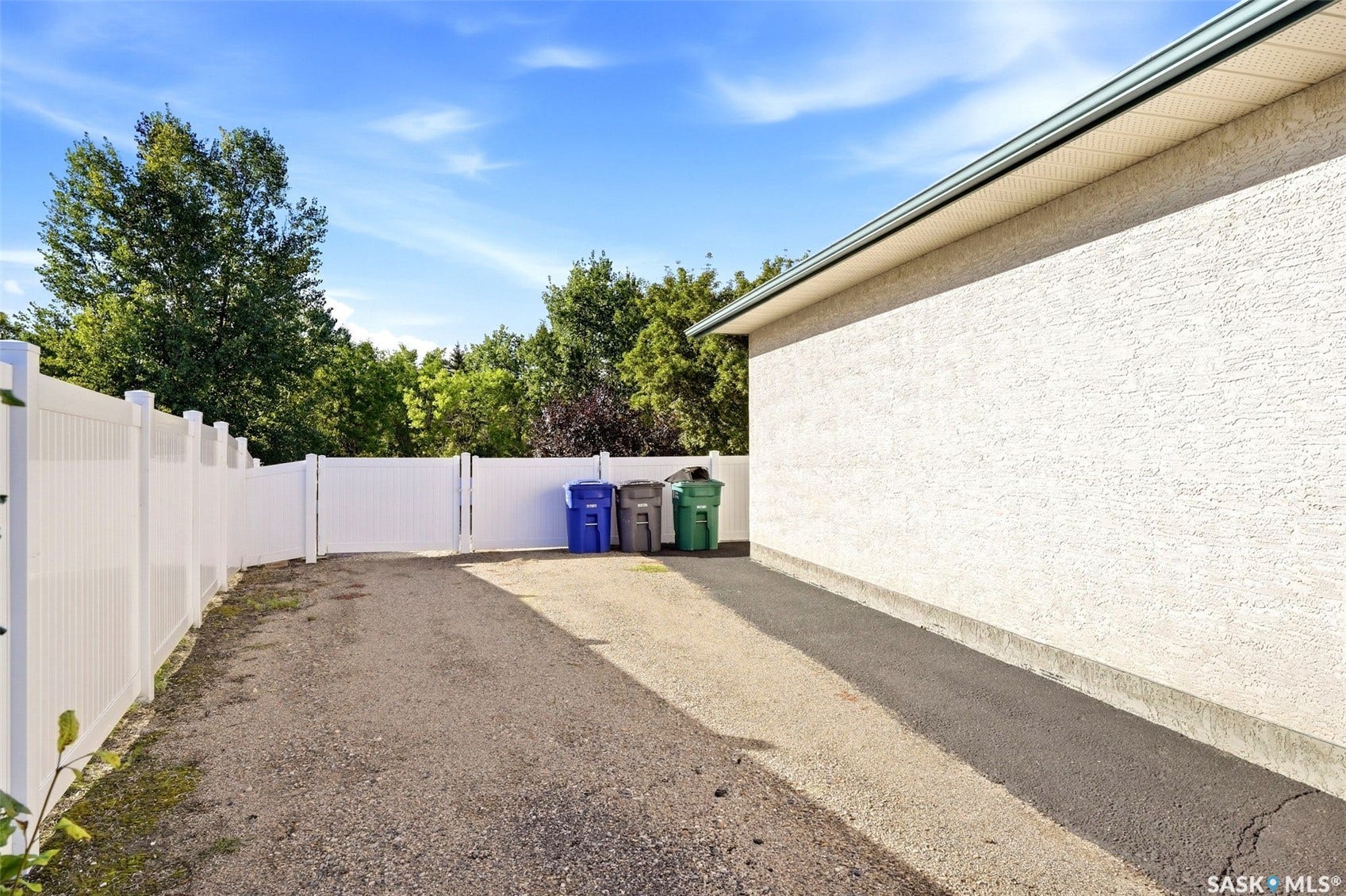 Stucco exterior with a soffit and gutter system, alongside a white privacy fence and gravel driveway