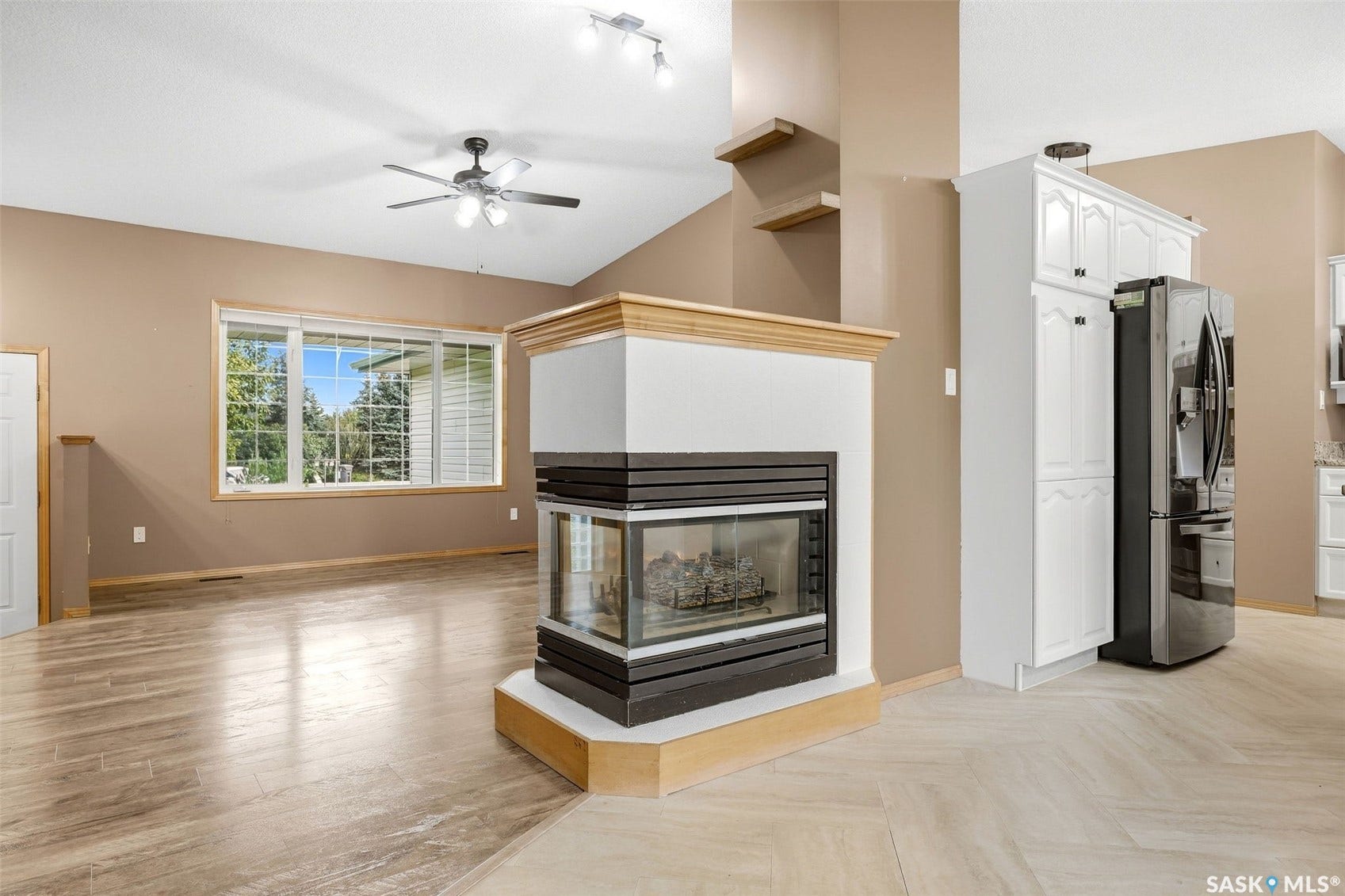Vaulted ceilings, a corner fireplace with a wood mantle, wood-finish flooring, a ceiling fan, and white cabinetry with a stainless steel refrigerator