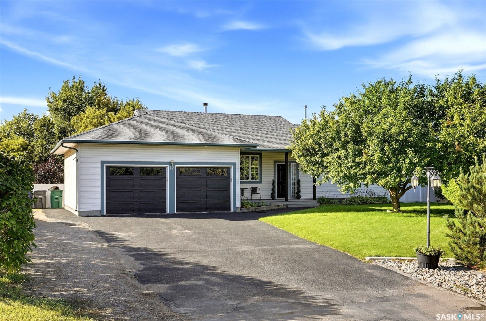 White siding exterior with a dark gray shingle roof, featuring two dark gray garage doors, a paved driveway, and established front yard landscaping with mature trees
