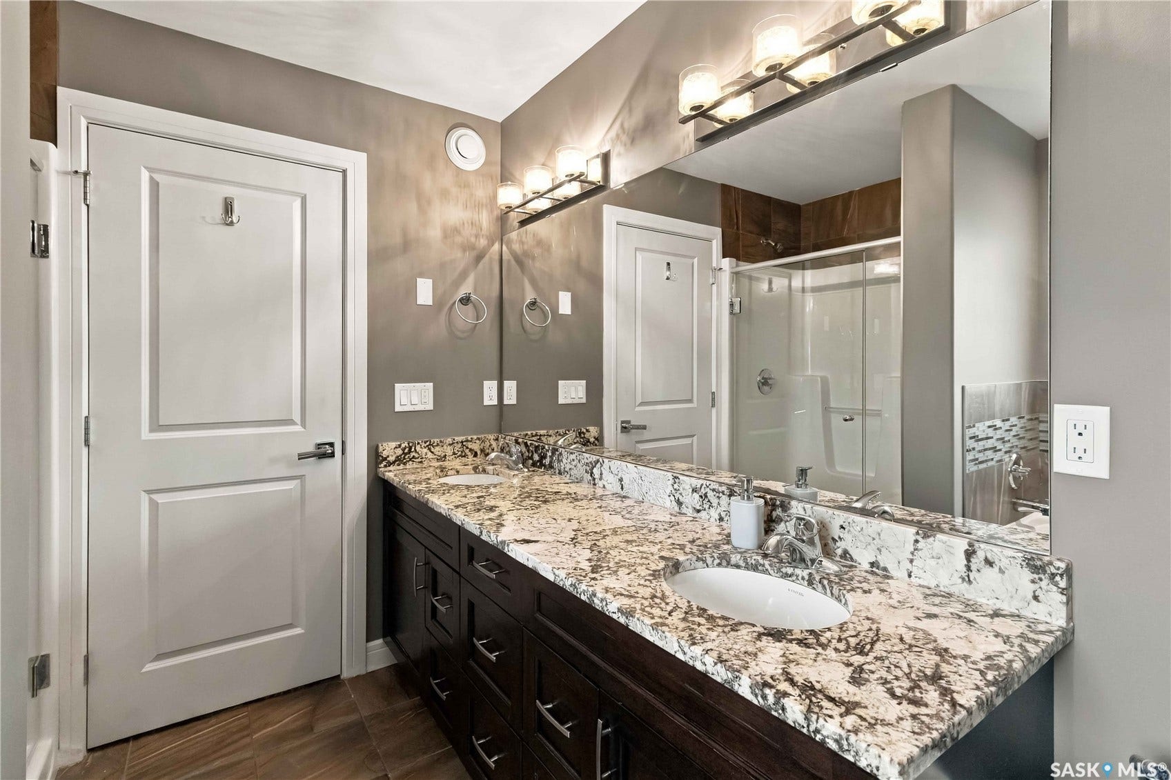 Dual undermount sinks with stone countertops, dark wood-finish cabinetry, wood-finish flooring, and a glass-enclosed shower
