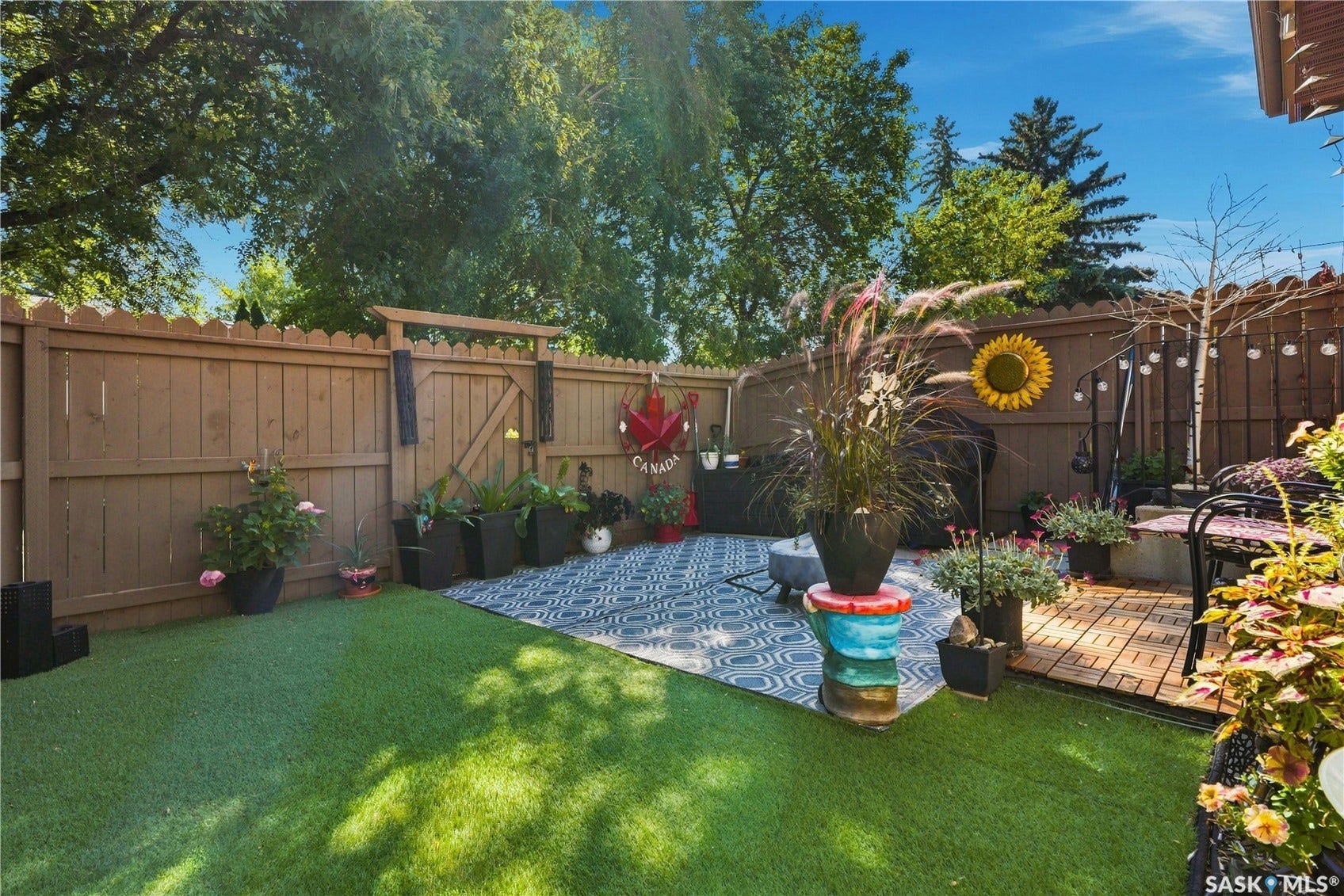 Fenced backyard featuring artificial turf, a patterned outdoor rug, a wood-finish deck area, and perimeter landscaping