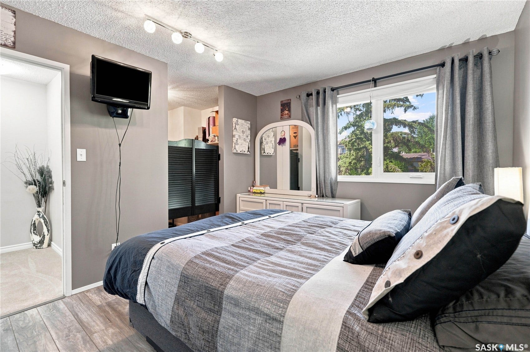 Wood-finish flooring throughout, window with exterior views, track lighting, wall-mounted television, and a built-in closet with louvered doors