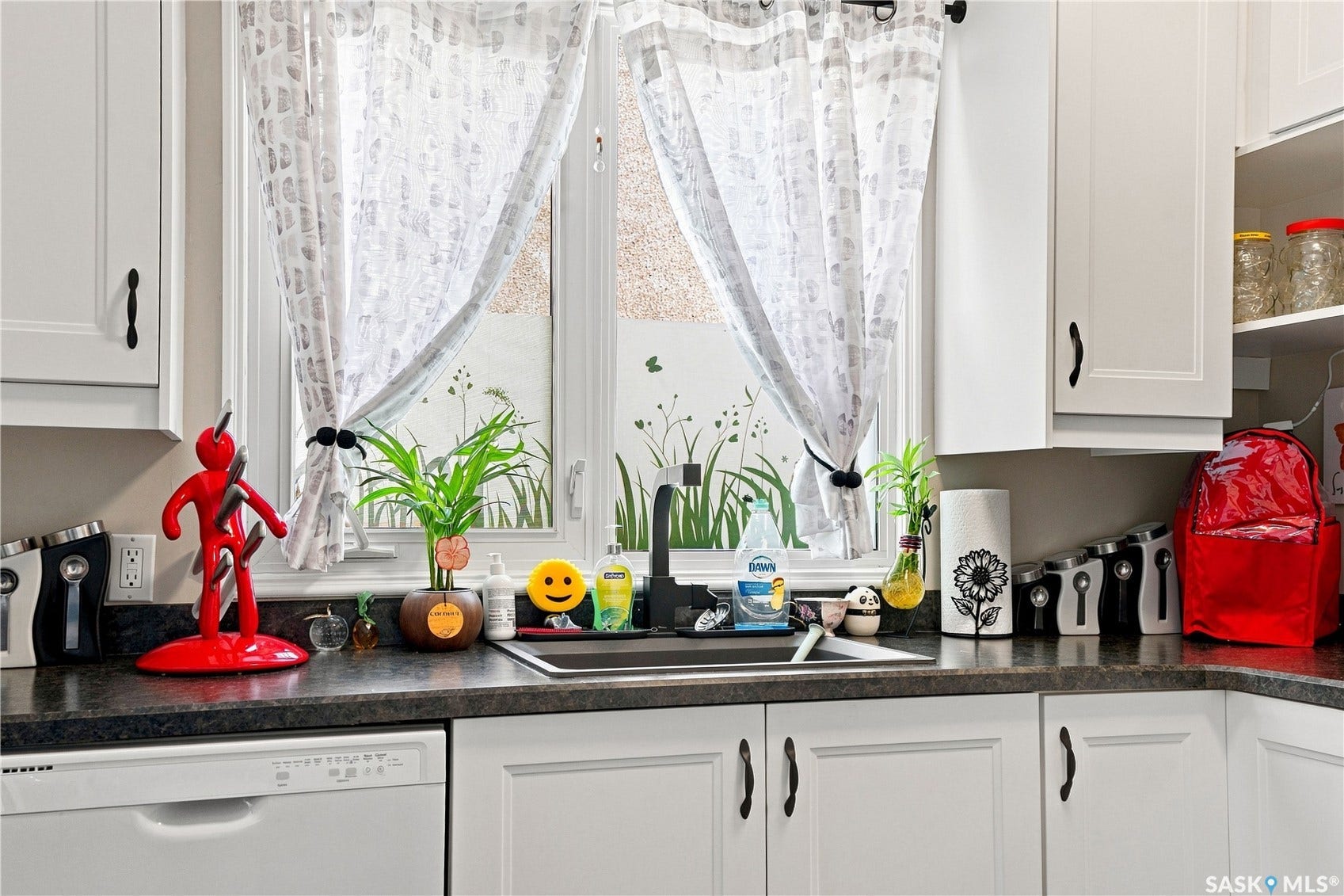 White cabinetry with dark hardware, dark countertop, stainless steel sink, and white built-in dishwasher