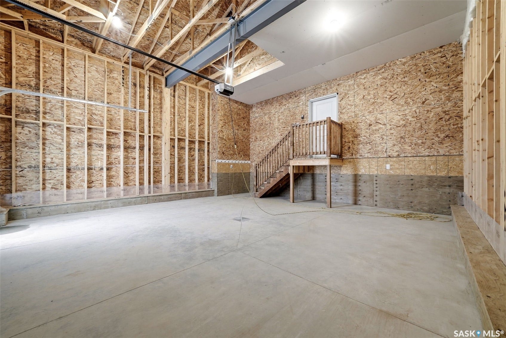 Concrete flooring, exposed wood framing, wood staircase with handrail, overhead lighting, and an overhead door mechanism