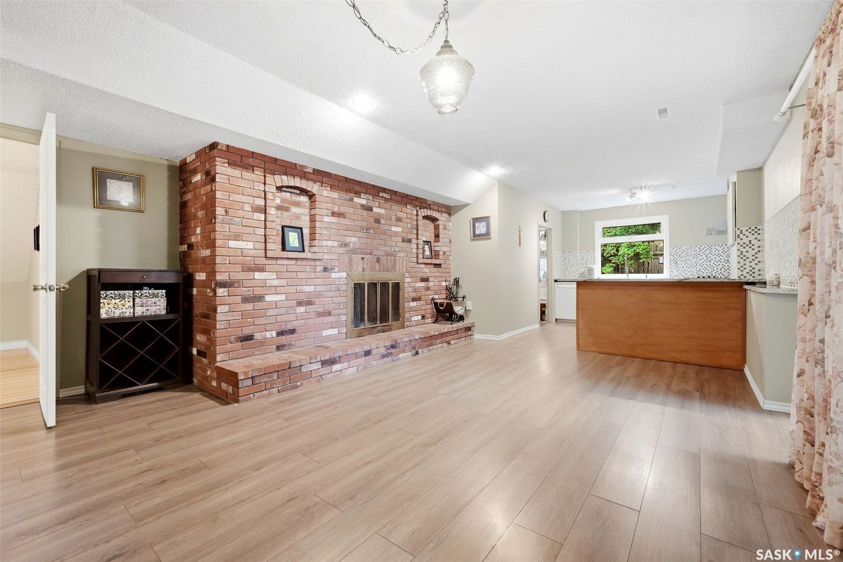 Exposed brick fireplace with hearth, wood-finish flooring, recessed lighting, kitchen island with wood-finish paneling, and a tiled kitchen backsplash